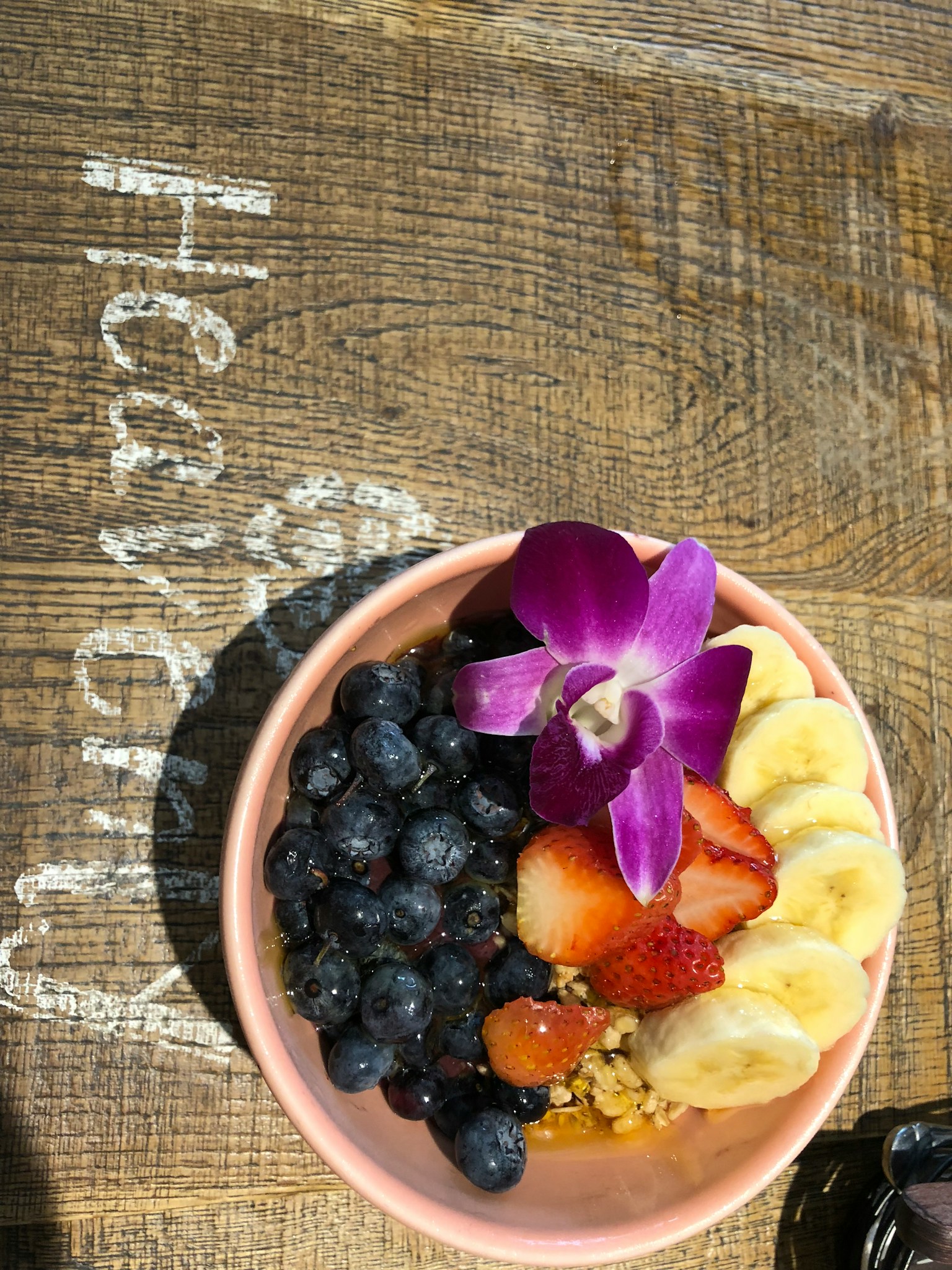Mangkok penuh dengan buah berwarna-warni termasuk blueberry pisang dan stroberi dihiasi dengan bunga ungu