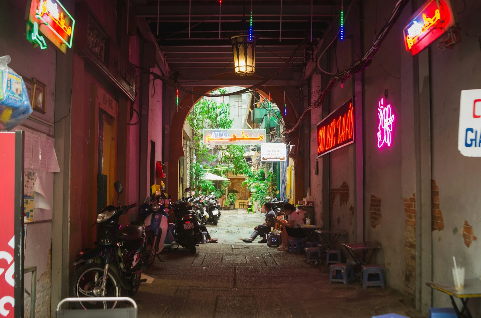 Schmale Gasse mit bunten Neonlichtern, geparkten Fahrrädern und sichtbarem Grün im Hintergrund