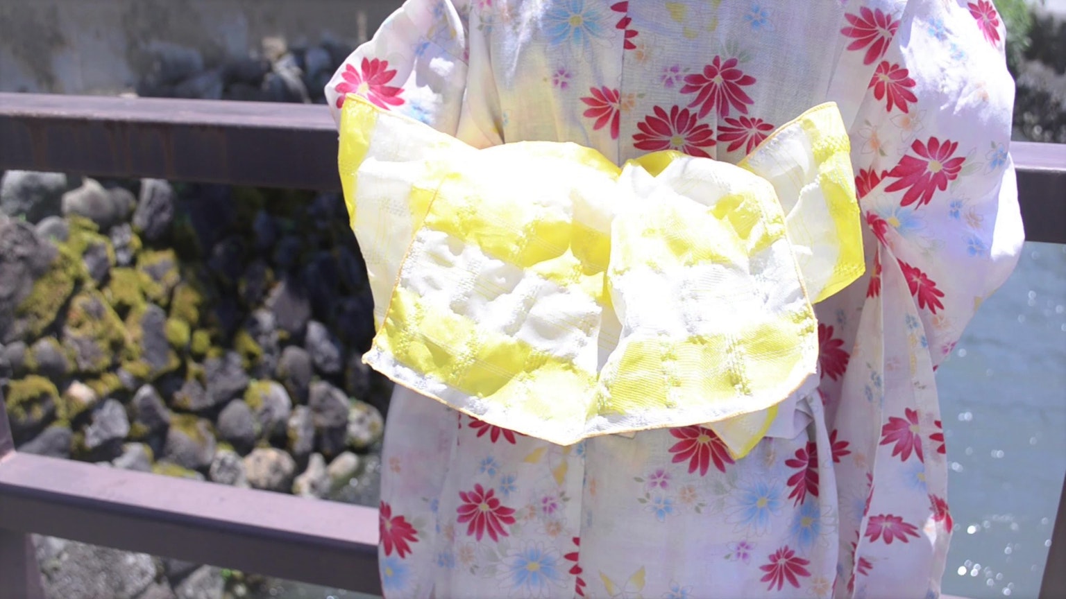Perempuan berpakaian kimono berbunga dengan obi kuning berdiri di dekat air