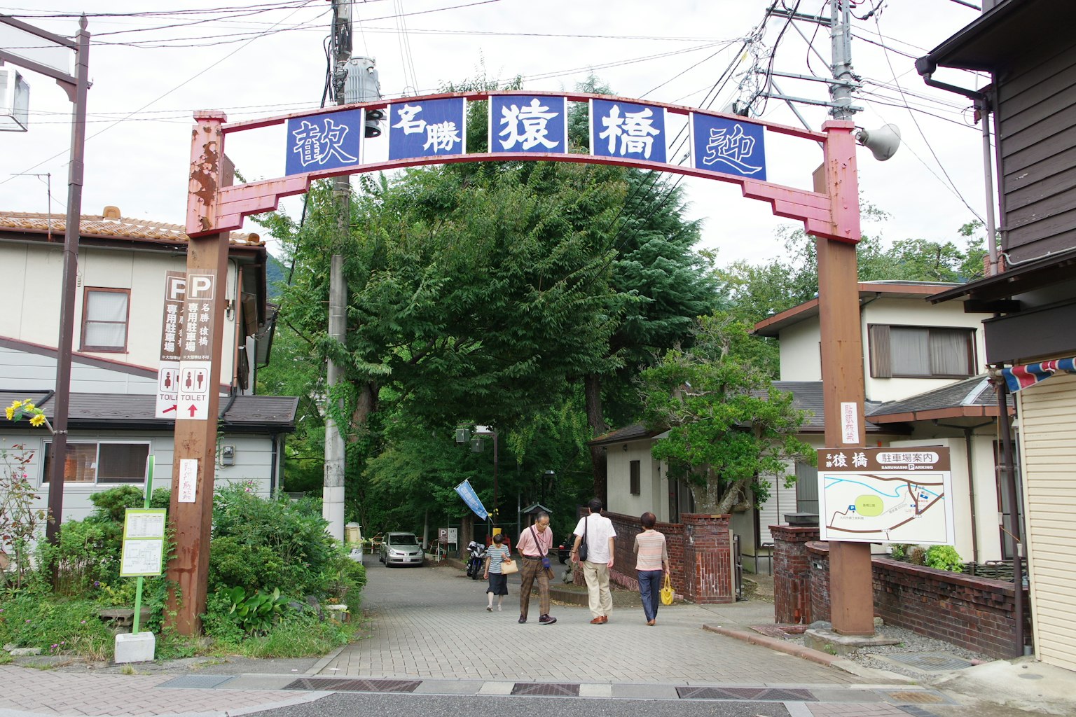 该图像展示了一个带有蓝色标志的拱门，通向一条绿树成荫的街道，有人正在走。