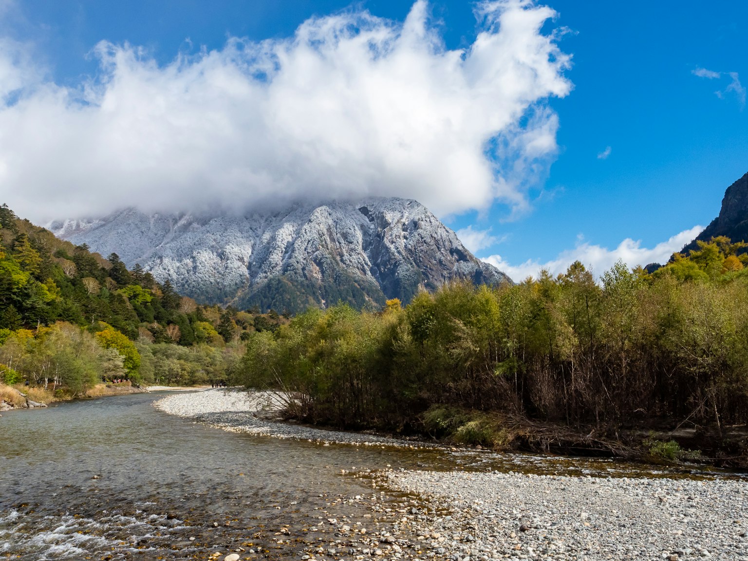 Scenic view of a mountain covered with clouds alongside a calm river and lush greenery