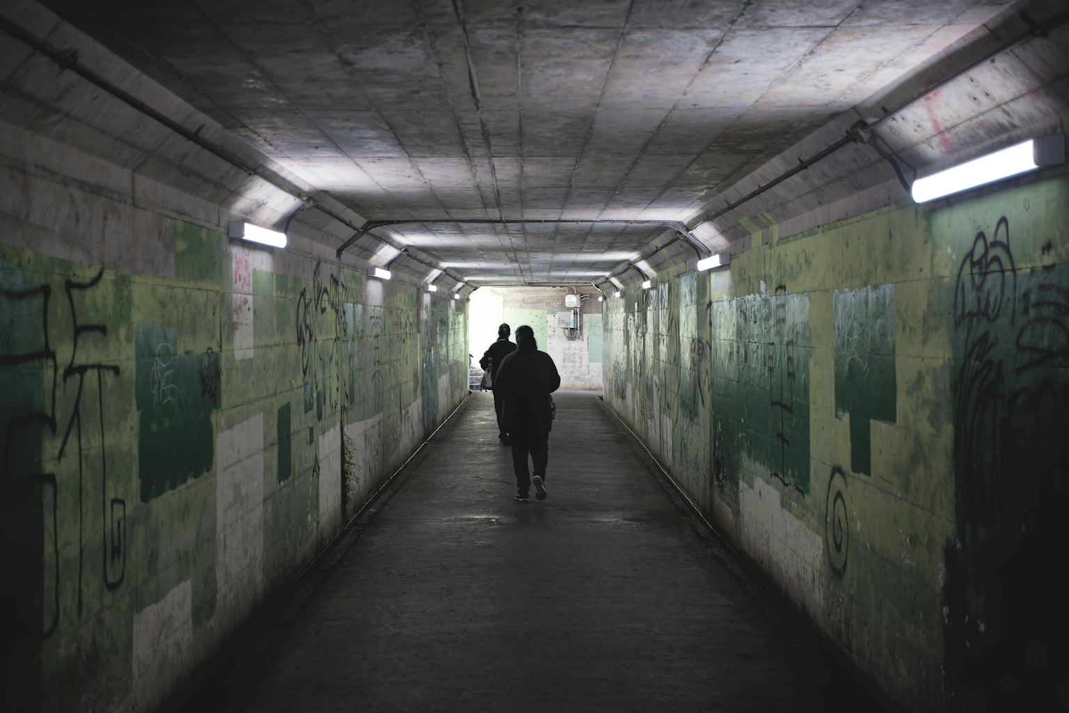 Dos figuras caminando en un túnel oscuro con paredes verdes y grafitis iluminados por luces tenues