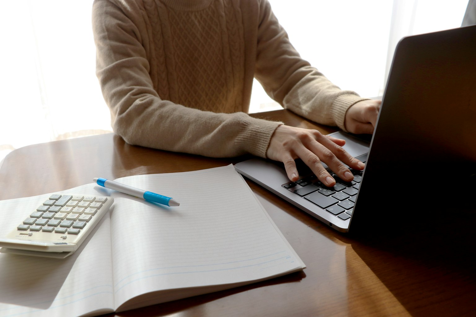 Nahaufnahme von Händen, die auf einem Laptop an einem Schreibtisch tippen, mit einem Taschenrechner, einem blauen Stift und einem leeren Notizbuch