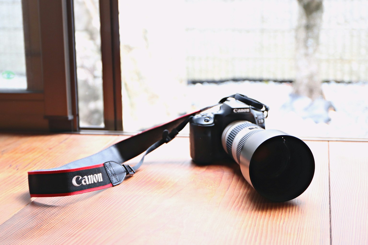 Kamera Canon dengan lensa diletakkan di atas meja kayu menghadap jendela