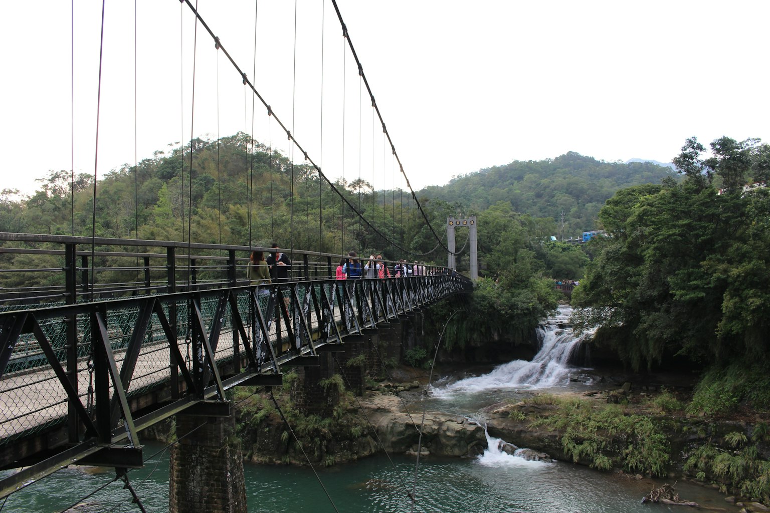 Orang-orang berjalan di jembatan gantung dengan latar belakang pegunungan hijau dan air terjun