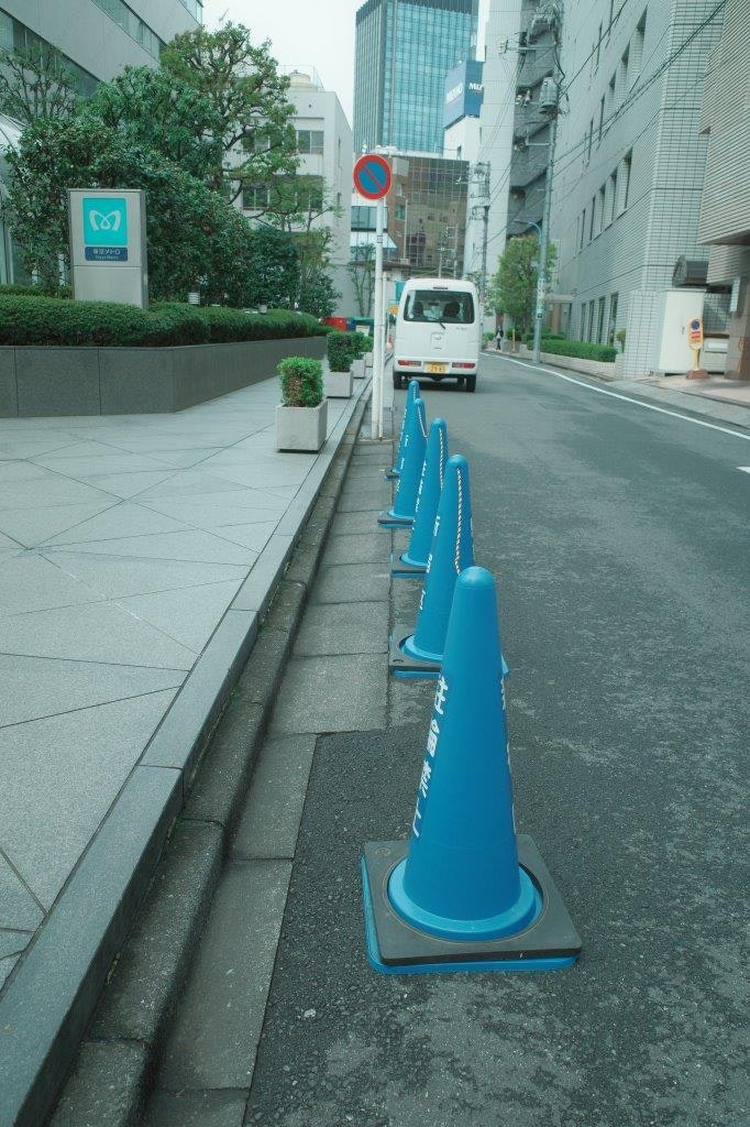 ビル街の歩道に並ぶ青いコーンと車道