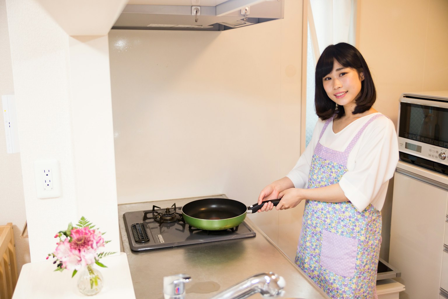 Joven mujer cocinando en la cocina sosteniendo una sartén verde usando un delantal de color claro