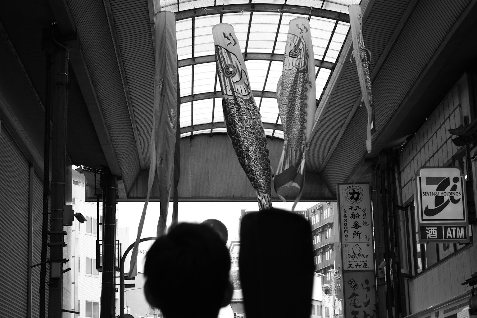Dos figuras en blanco y negro bajo un techo con banderas de koi ondeando
