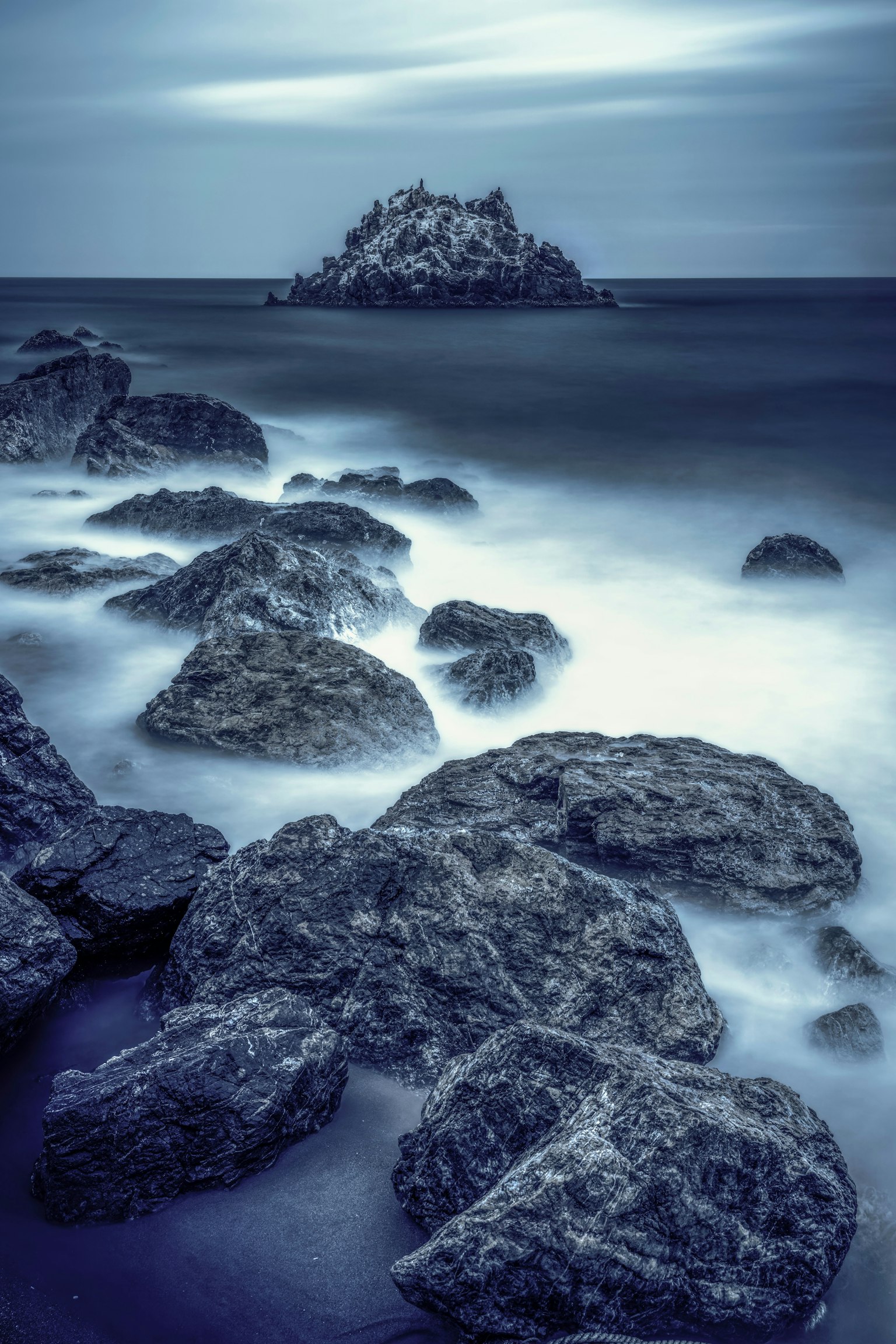 Coastal scene with scattered rocks and a distant island in blue tones