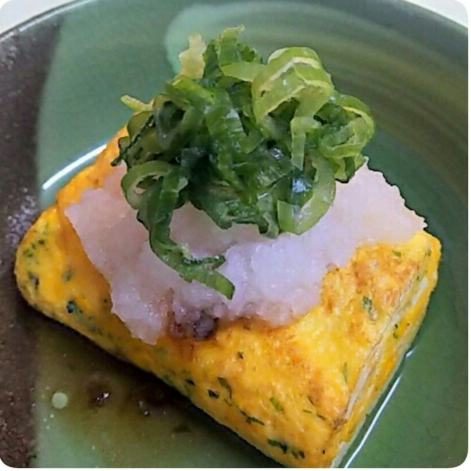 Ein Stück japanisches Omelett, garniert mit geriebenem Daikon und gehackten Frühlingszwiebeln auf einem grünen Teller