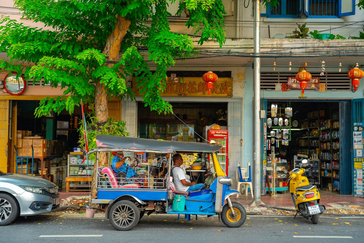 Un tuk-tuk blu parcheggiato davanti a un negozio con alberi verdi e lanterne