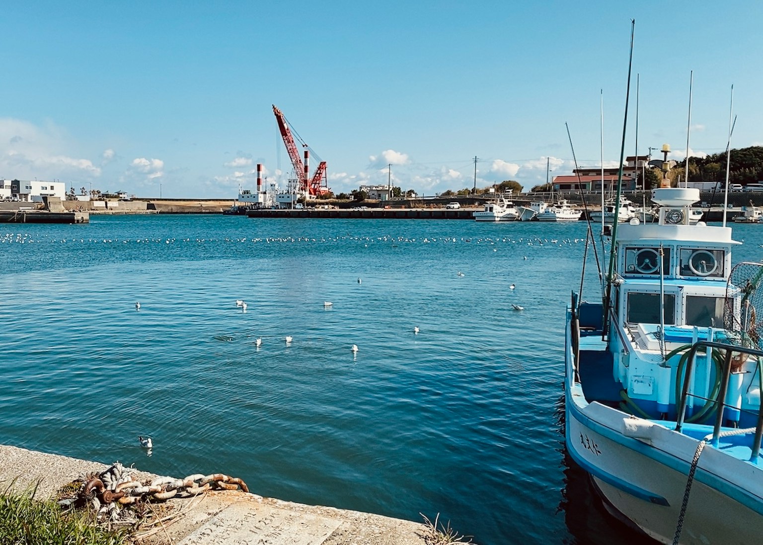 A blue fishing boat docked in a calm harbor with a construction crane in the background
