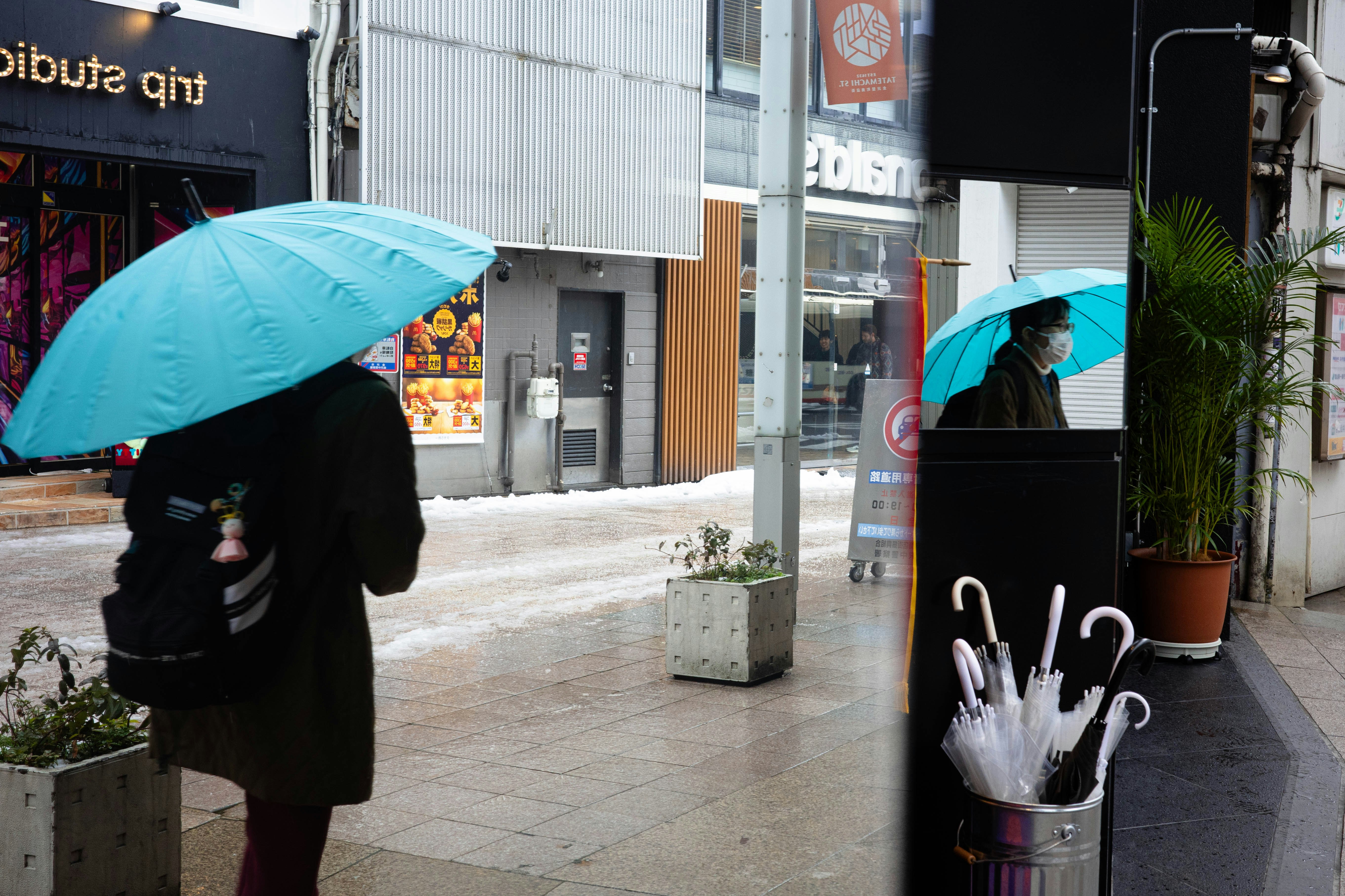 青い傘をさした人が並ぶ雨の路地の店先