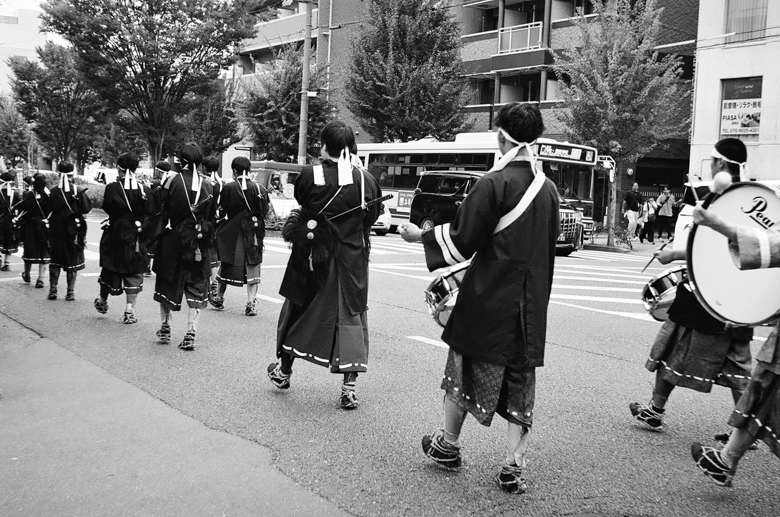 Eine Prozession von Menschen in traditioneller Kleidung, die eine Straße entlanggehen, mit einem Bus im Hintergrund in Schwarzweiß