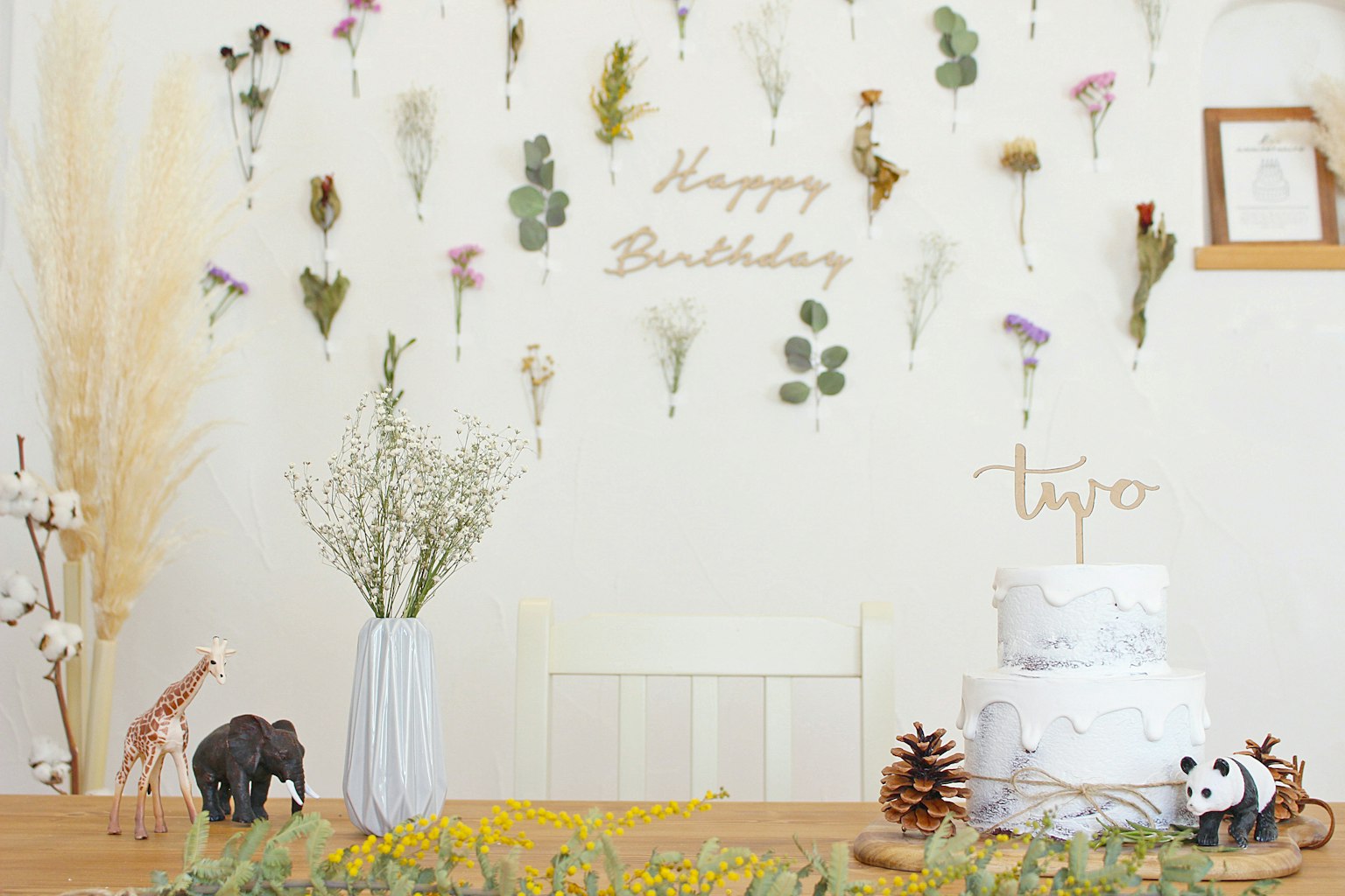 A birthday decoration table featuring dried flowers on the wall and a cake with small animal figurines