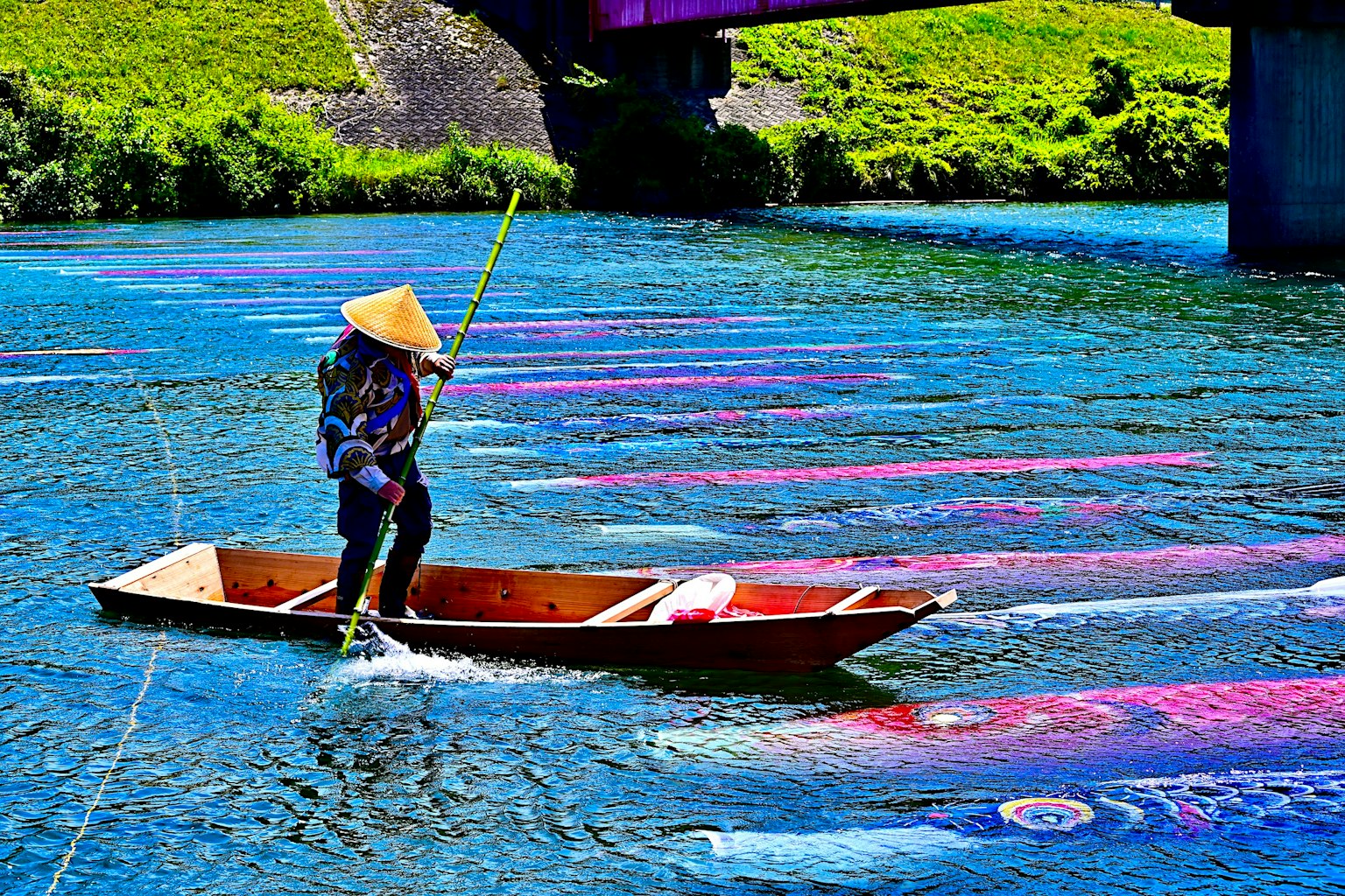 บุคคลที่สวมหมวกทรงกรวยพายเรือในแม่น้ำที่มีผ้าหลากสีลอยอยู่