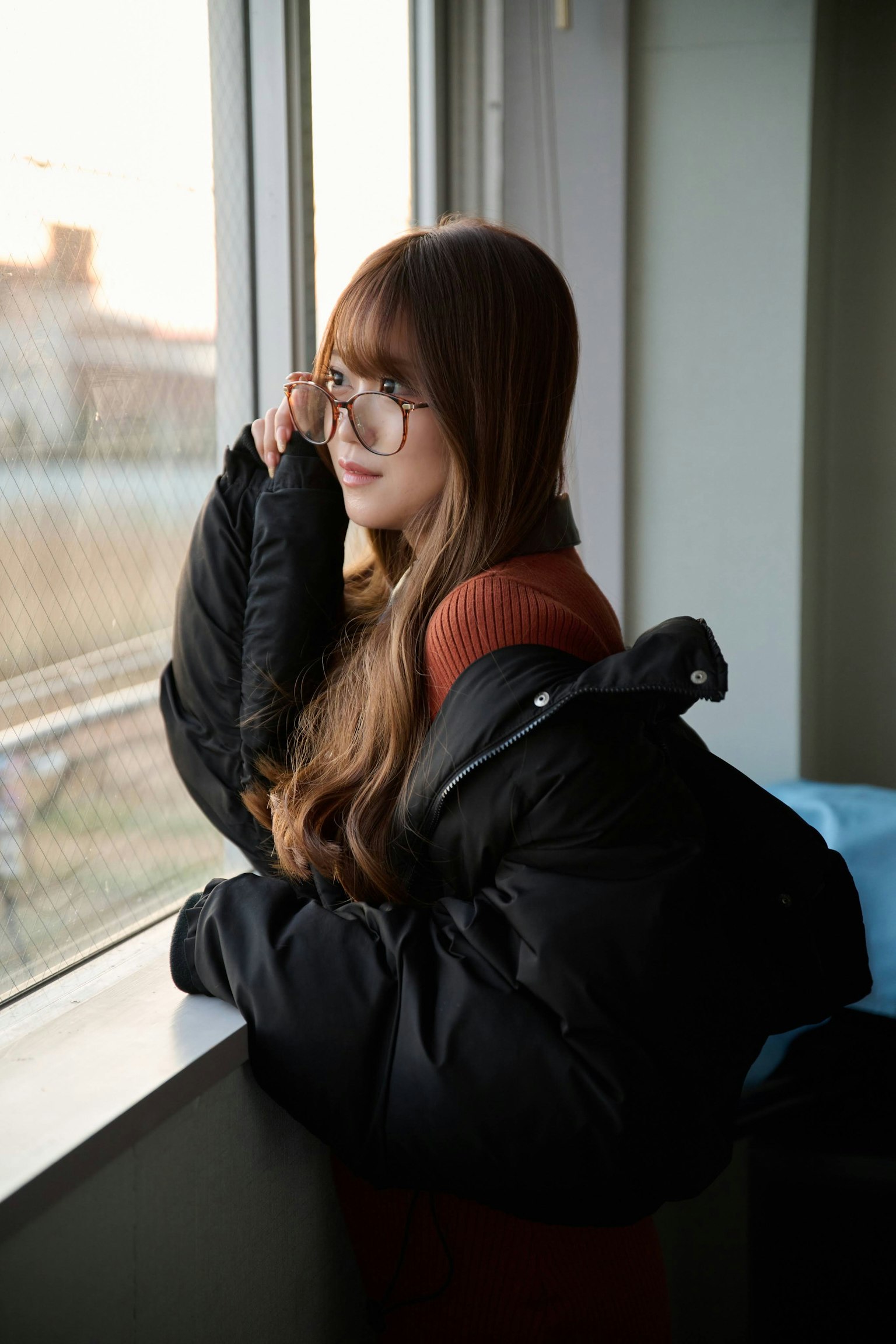 Wanita berdiri di dekat jendela dengan rambut panjang mengenakan jaket puffer hitam dan kacamata