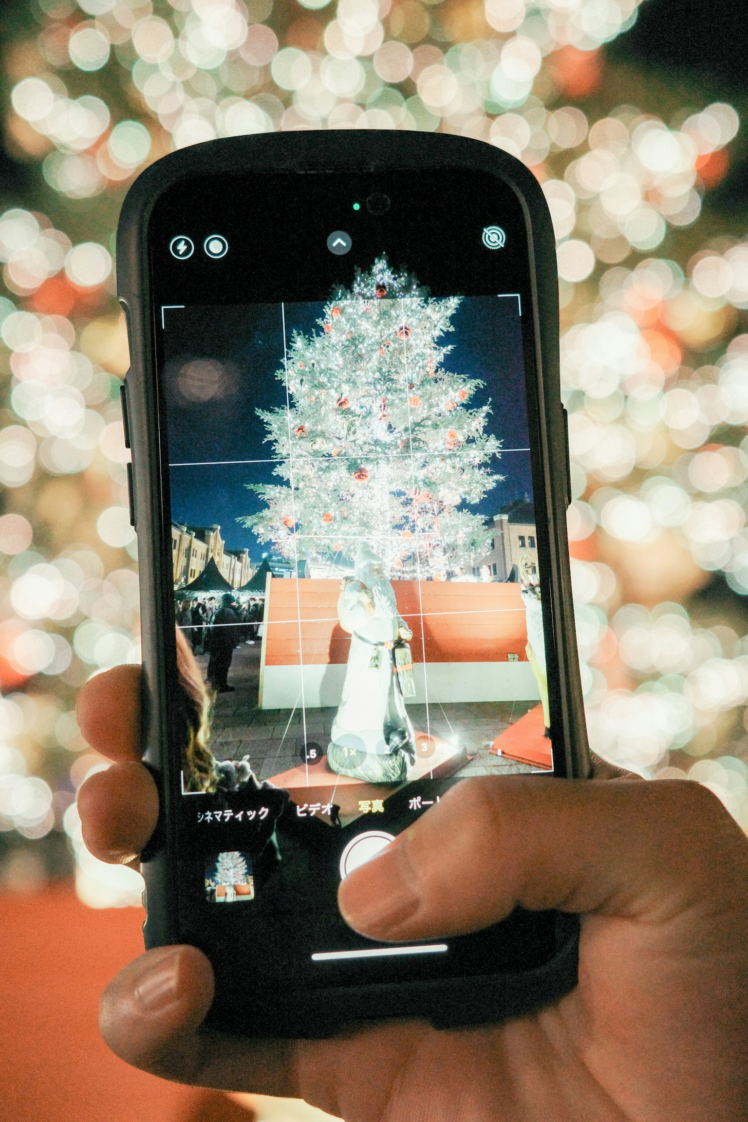 手に持たれたスマートフォンで撮影されたクリスマスツリーの写真背景にはぼんやりとした光の玉がある