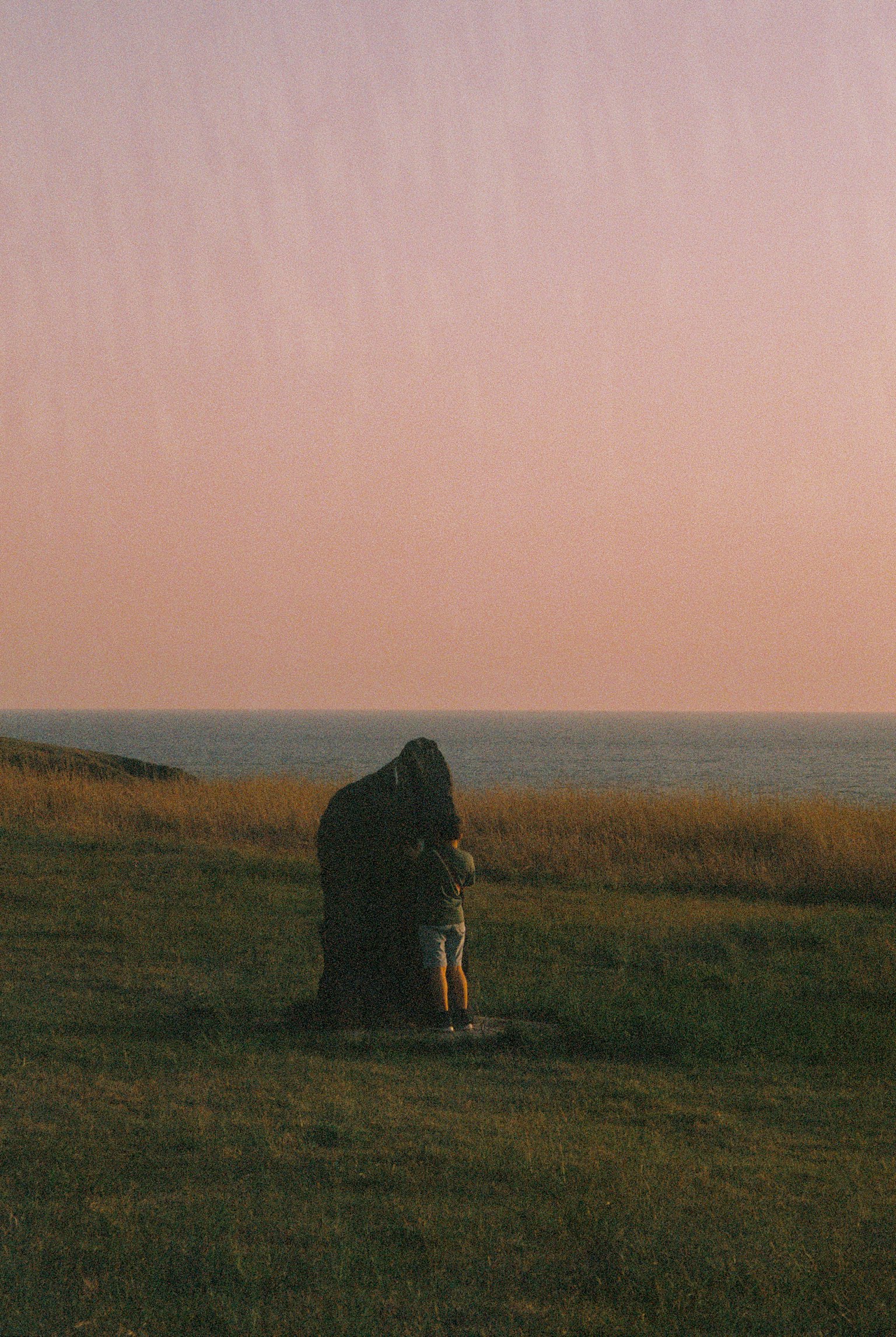 Người đứng bên một tảng đá lớn trong cánh đồng cỏ với hoàng hôn trên đại dương