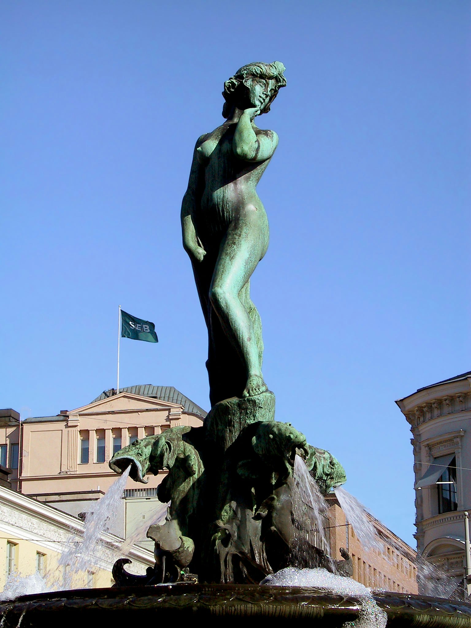Una statua verde si erge al centro di una fontana con cielo blu e edifici sullo sfondo