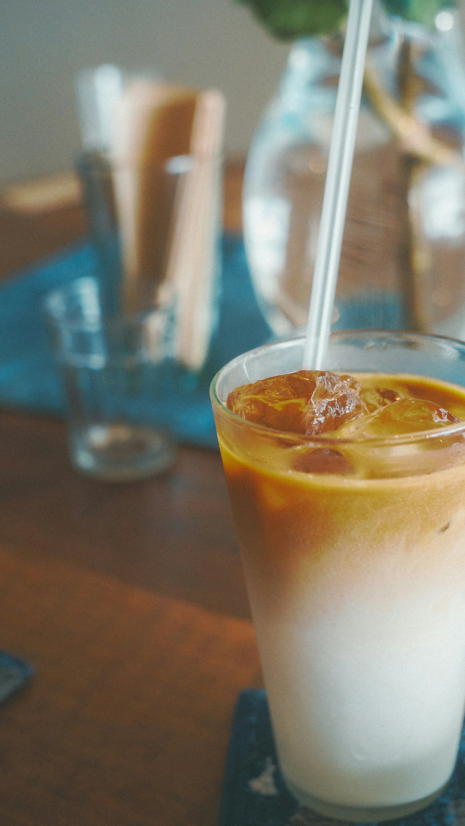 A glass of iced coffee on a table with a transparent water pitcher and cutlery in the background