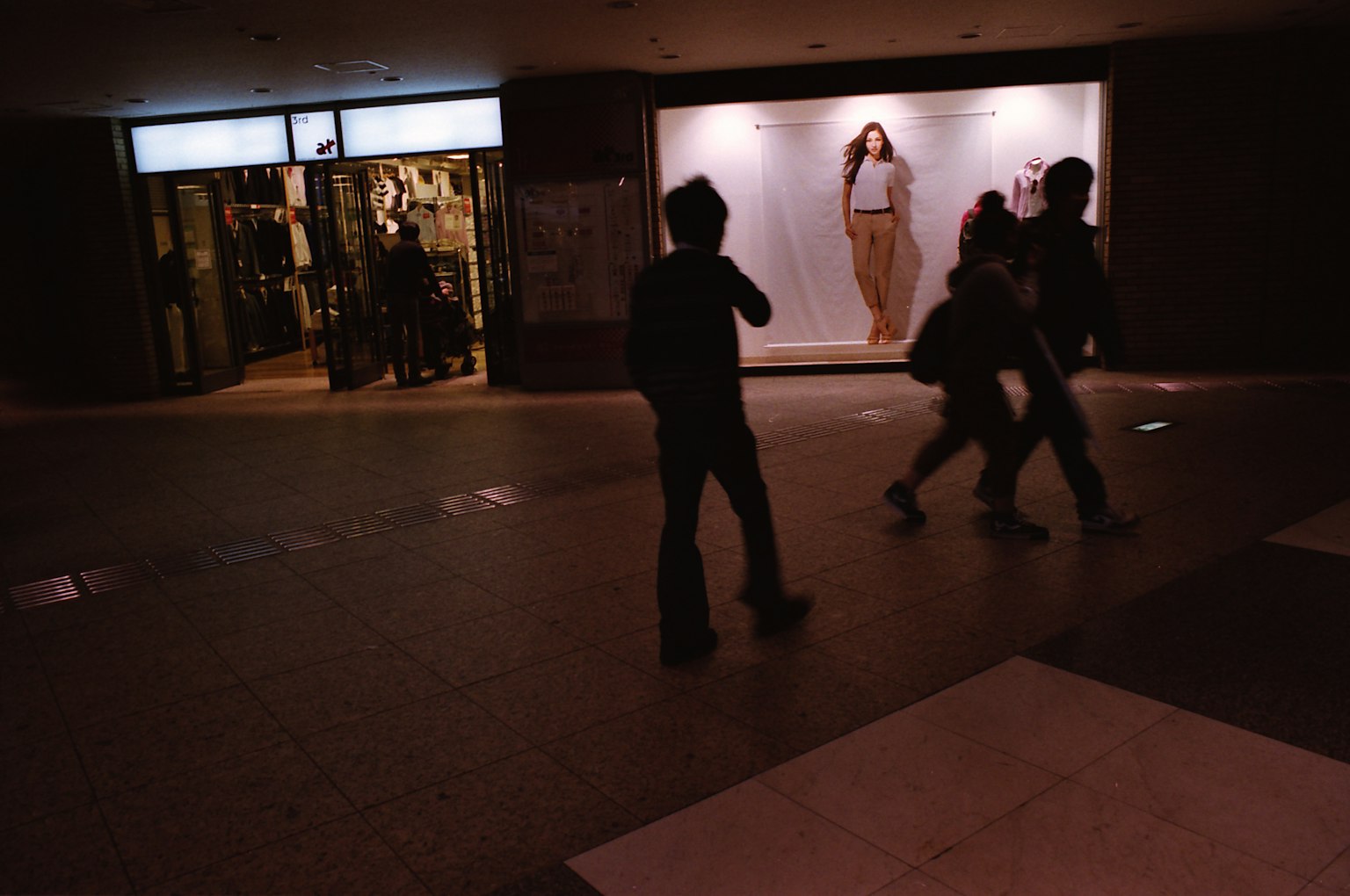 Personas caminando en un pasillo de centro comercial poco iluminado con un modelo de moda en la vitrina