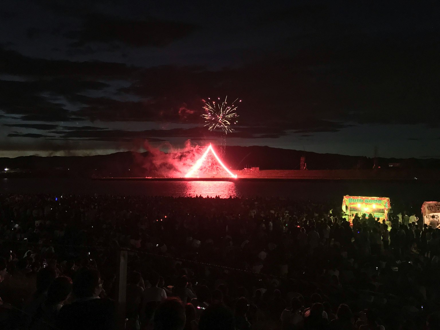 Scène de festival avec des feux d'artifice formant une forme triangulaire rouge dans le ciel nocturne