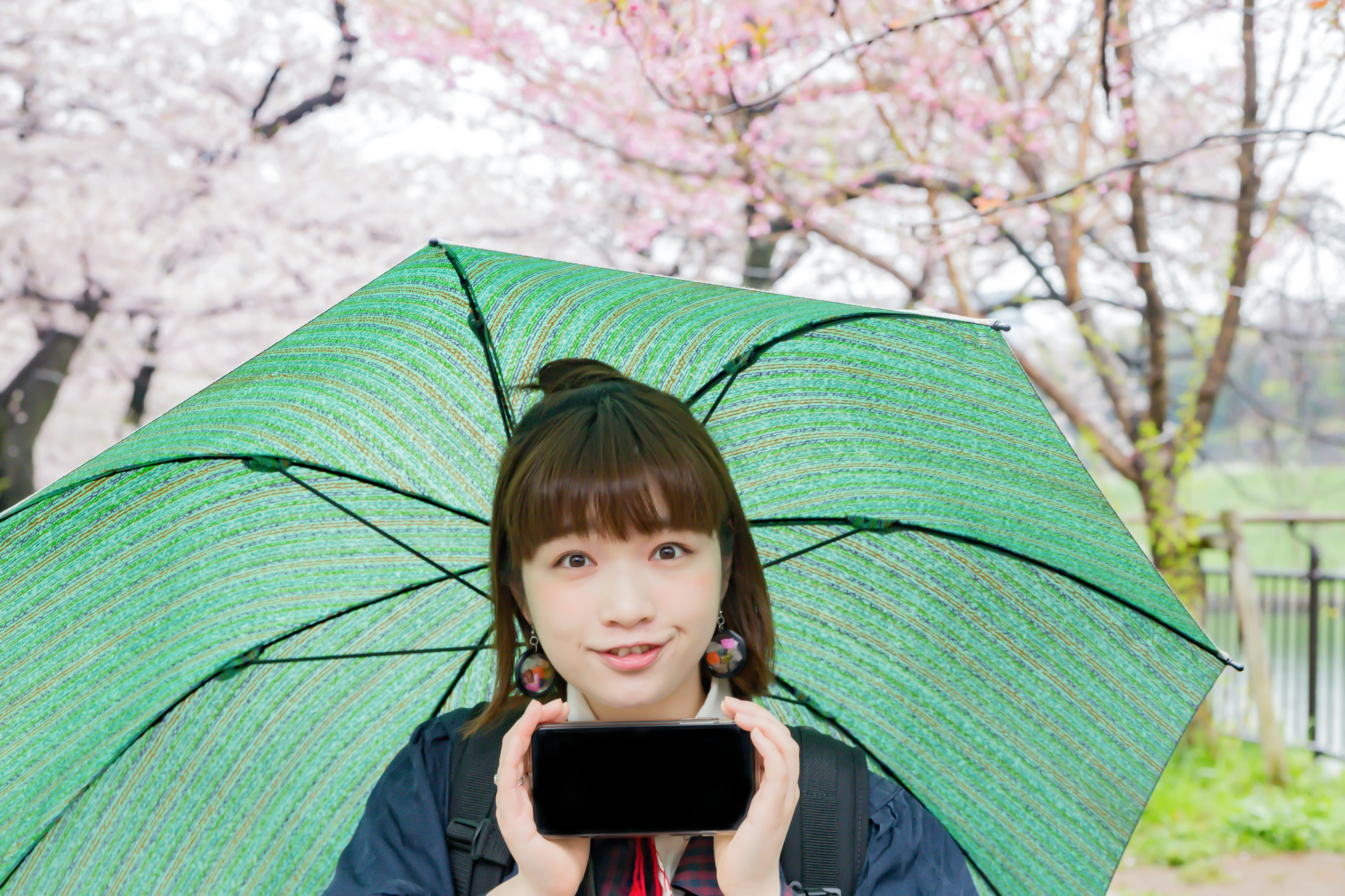 櫻花下撐綠色傘、手持手機的女性