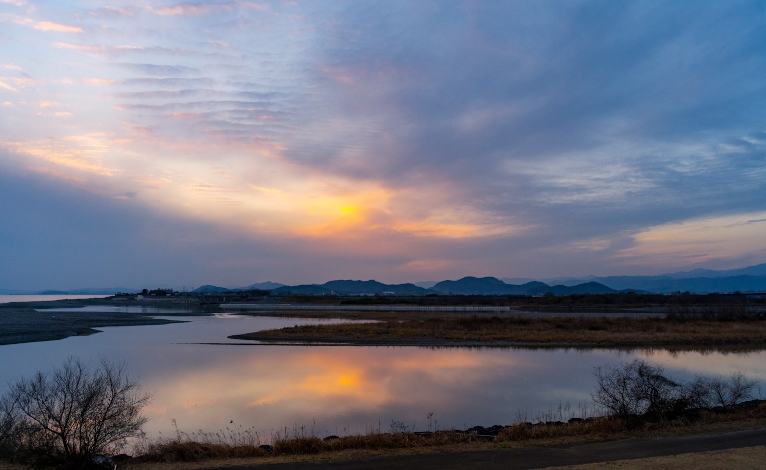 Tranquil sunset landscape with river reflection, blue sky and orange sun, silhouette of mountains