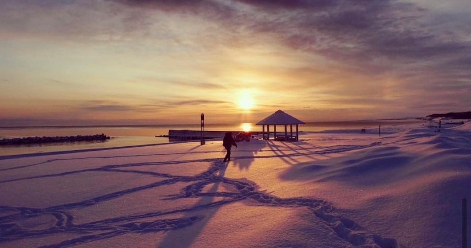 Person walking on snowy beach at sunset with pavilion