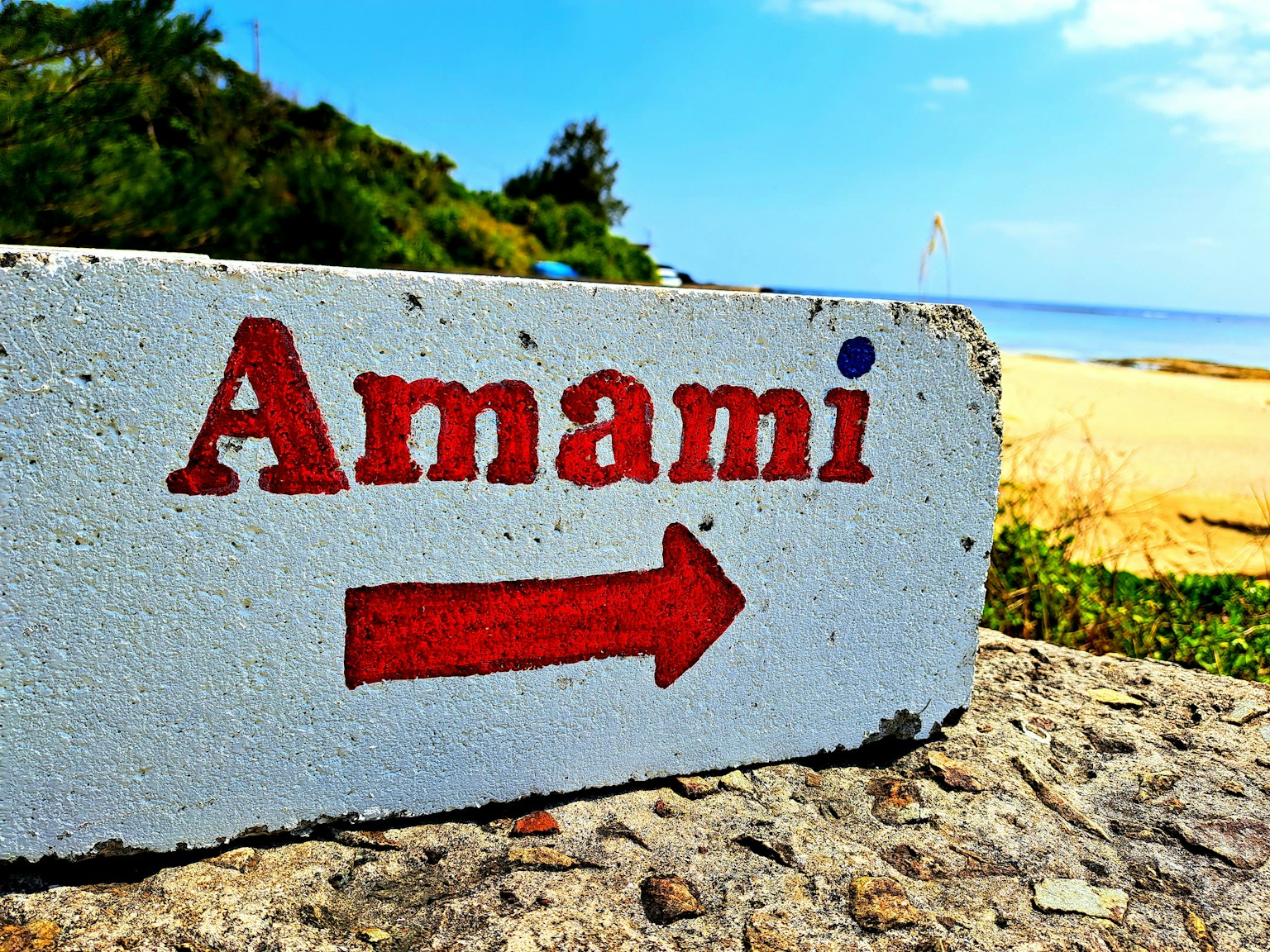 Cartel que indica dirección a Amami con cielo azul y playa de fondo