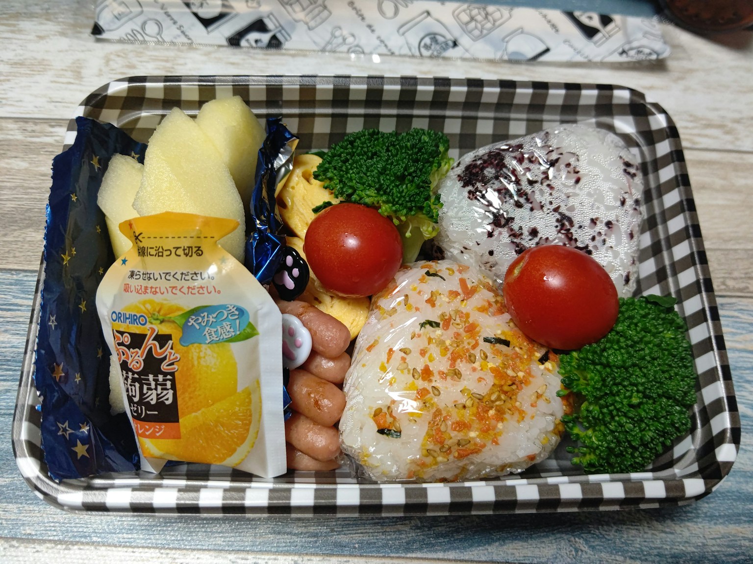 Kotak bento dengan bola nasi, brokoli, tomat ceri, sosis, pisang, dan minuman