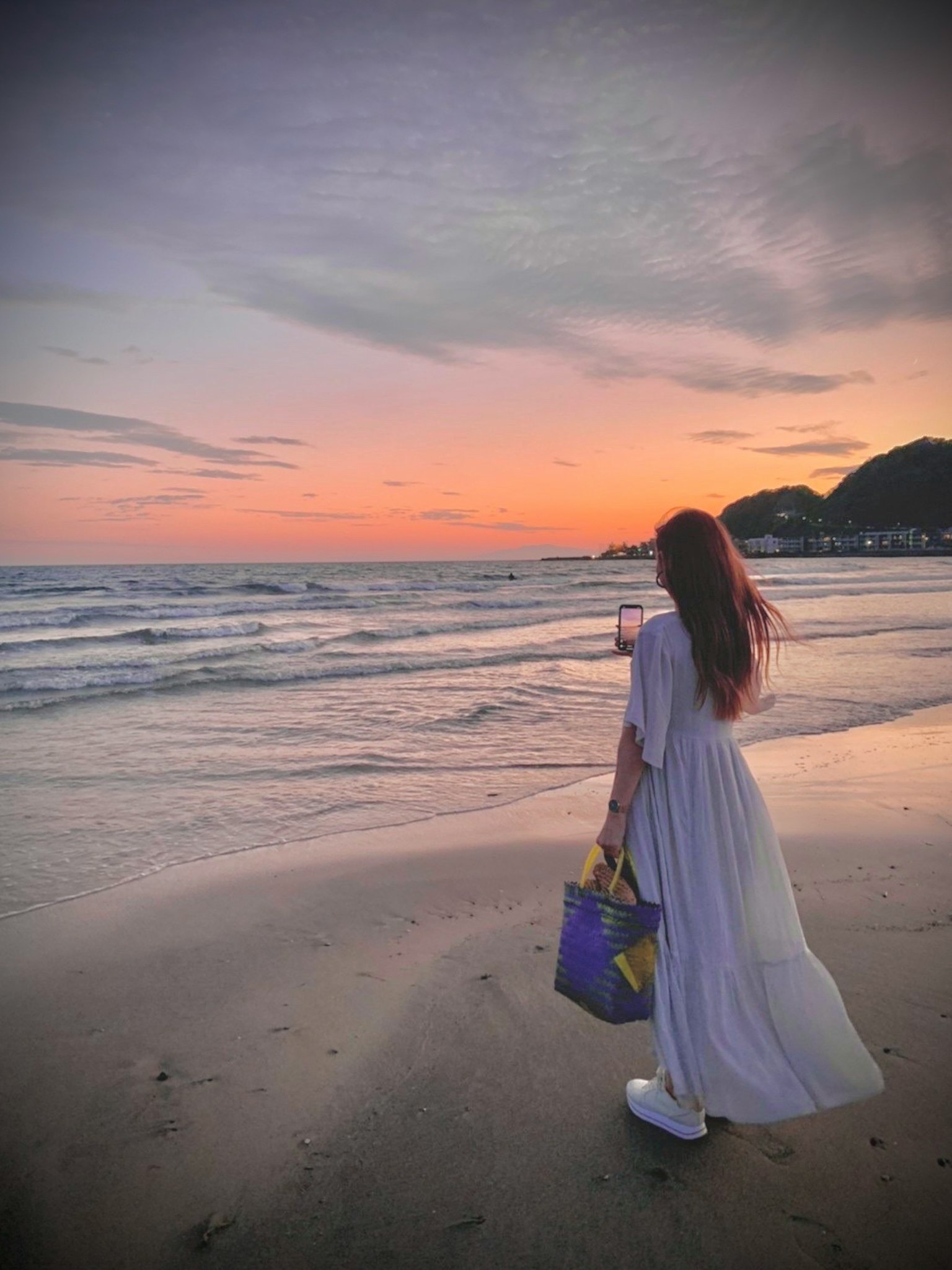Seorang wanita berpakaian putih berdiri di pantai dengan matahari terbenam di latar belakang