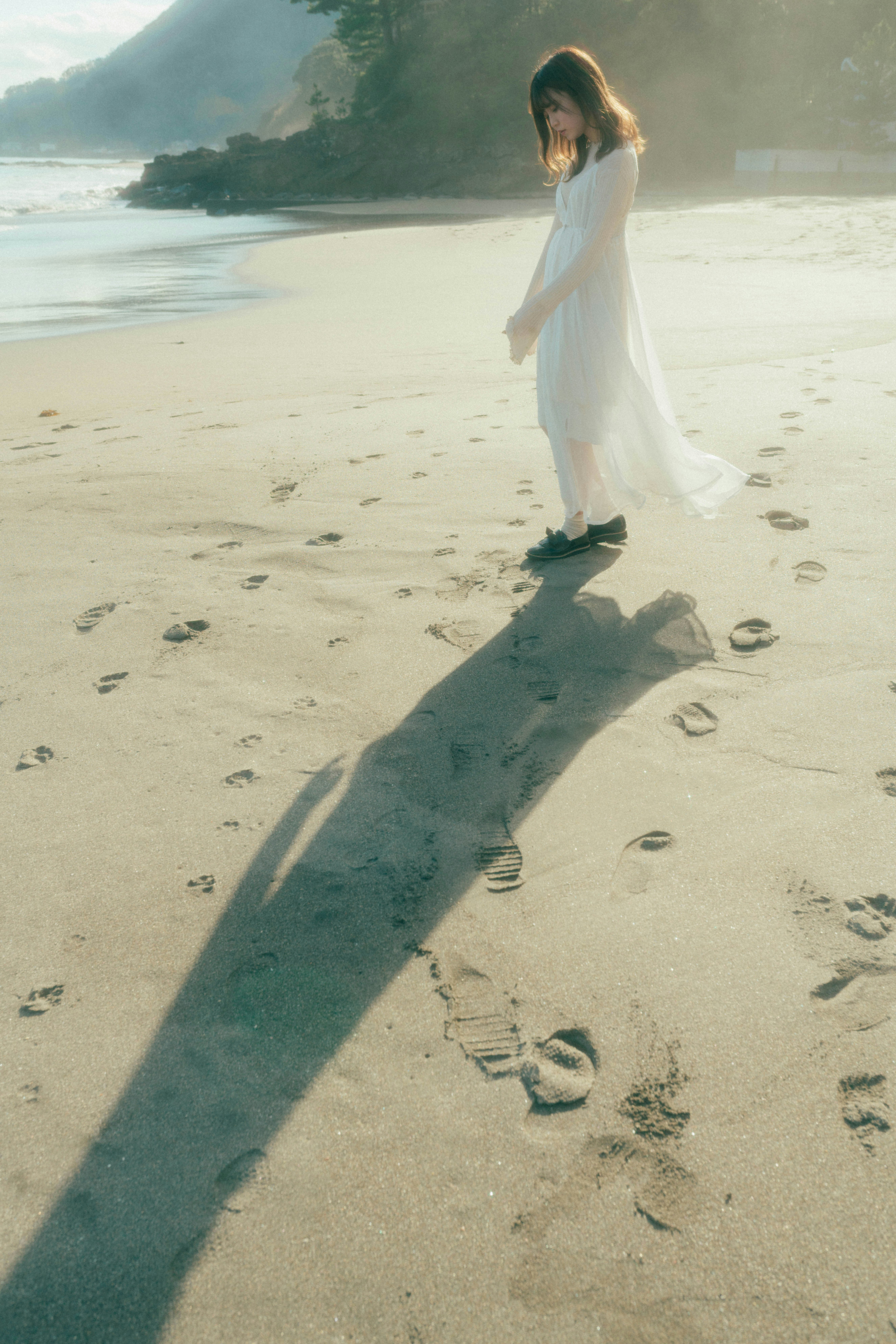砂浜を歩く白いワンピースの女性と波打ち際の海景