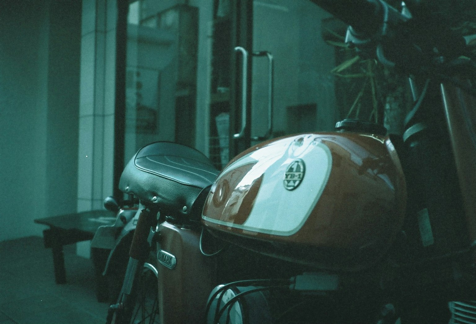 Gambar samping sepeda motor dengan tangki oranye bangunan kaca terlihat di latar belakang