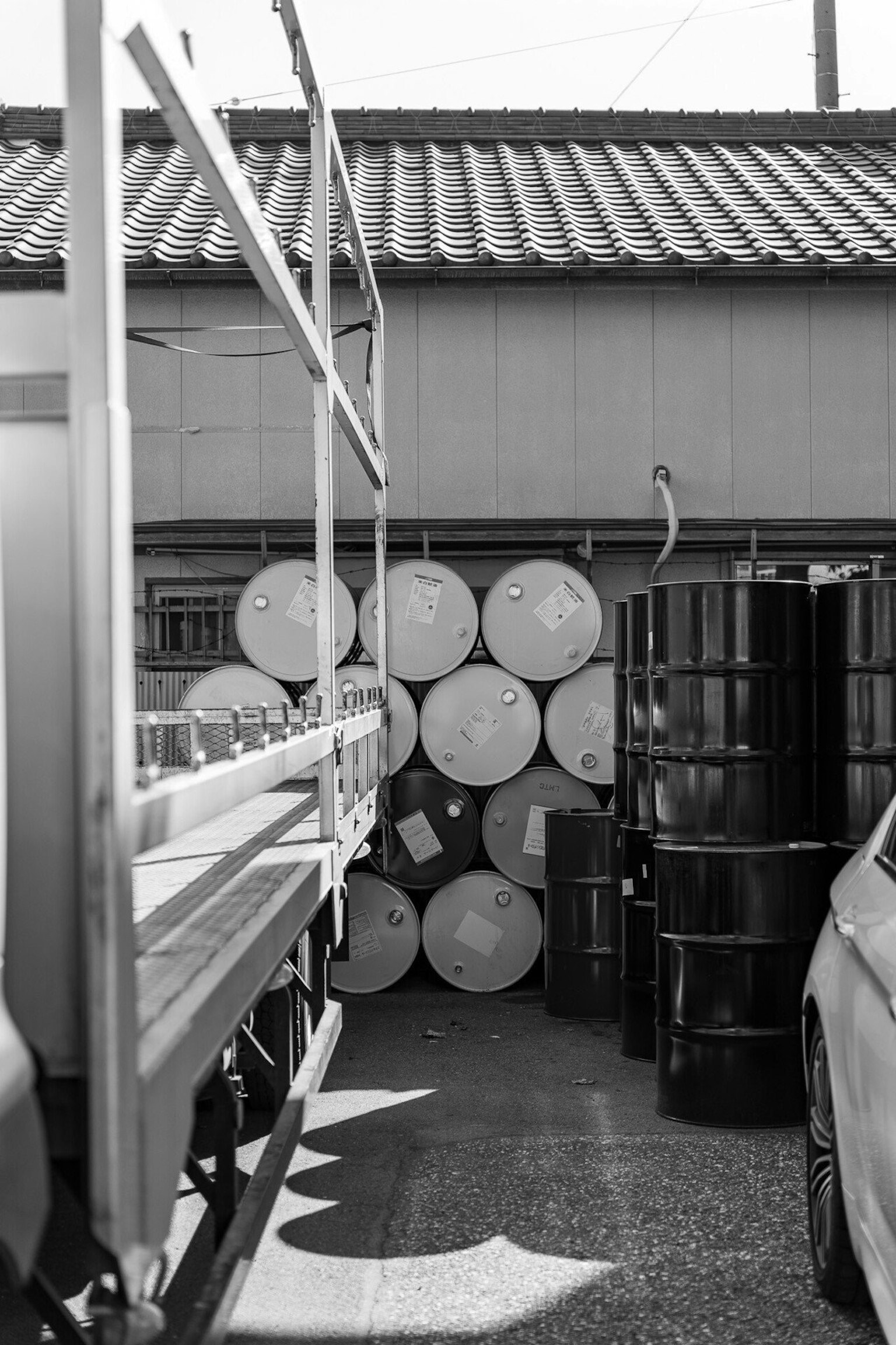 Black and white image of stacked barrels behind a warehouse with part of a truck