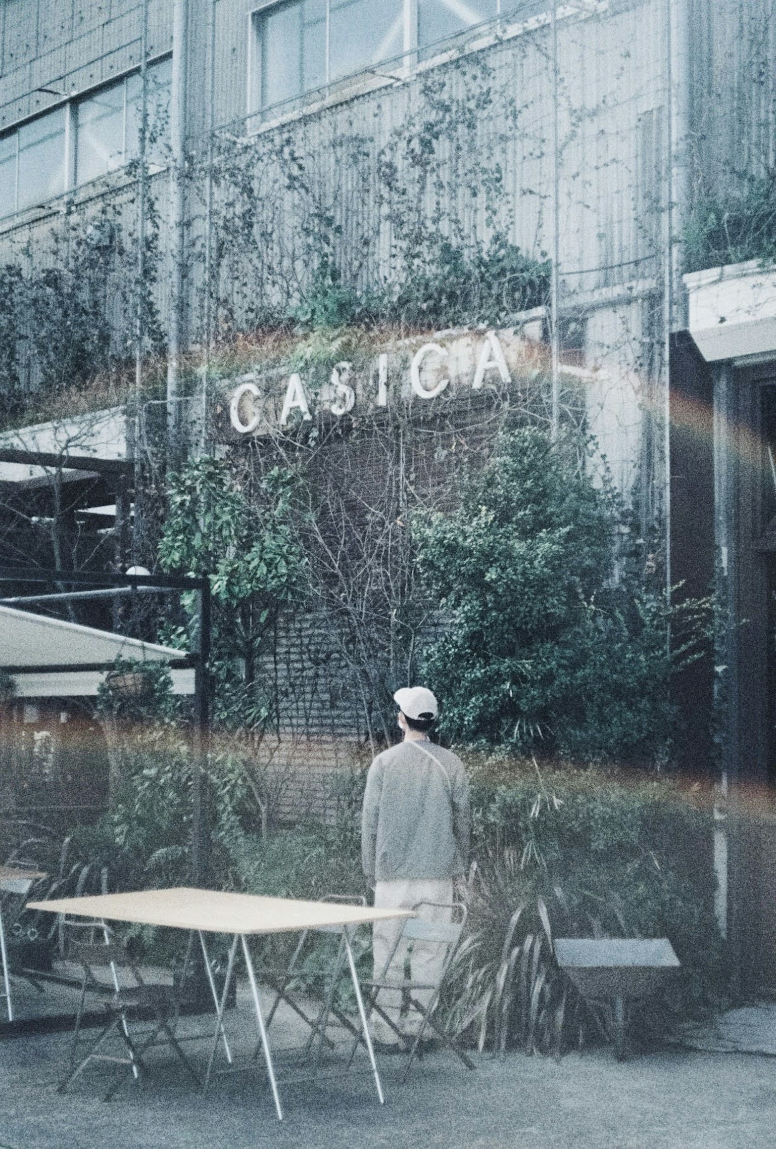 ด้านนอกของคาเฟ่ที่เงียบสงบล้อมรอบด้วยพืชสีเขียวและมีคนยืนอยู่