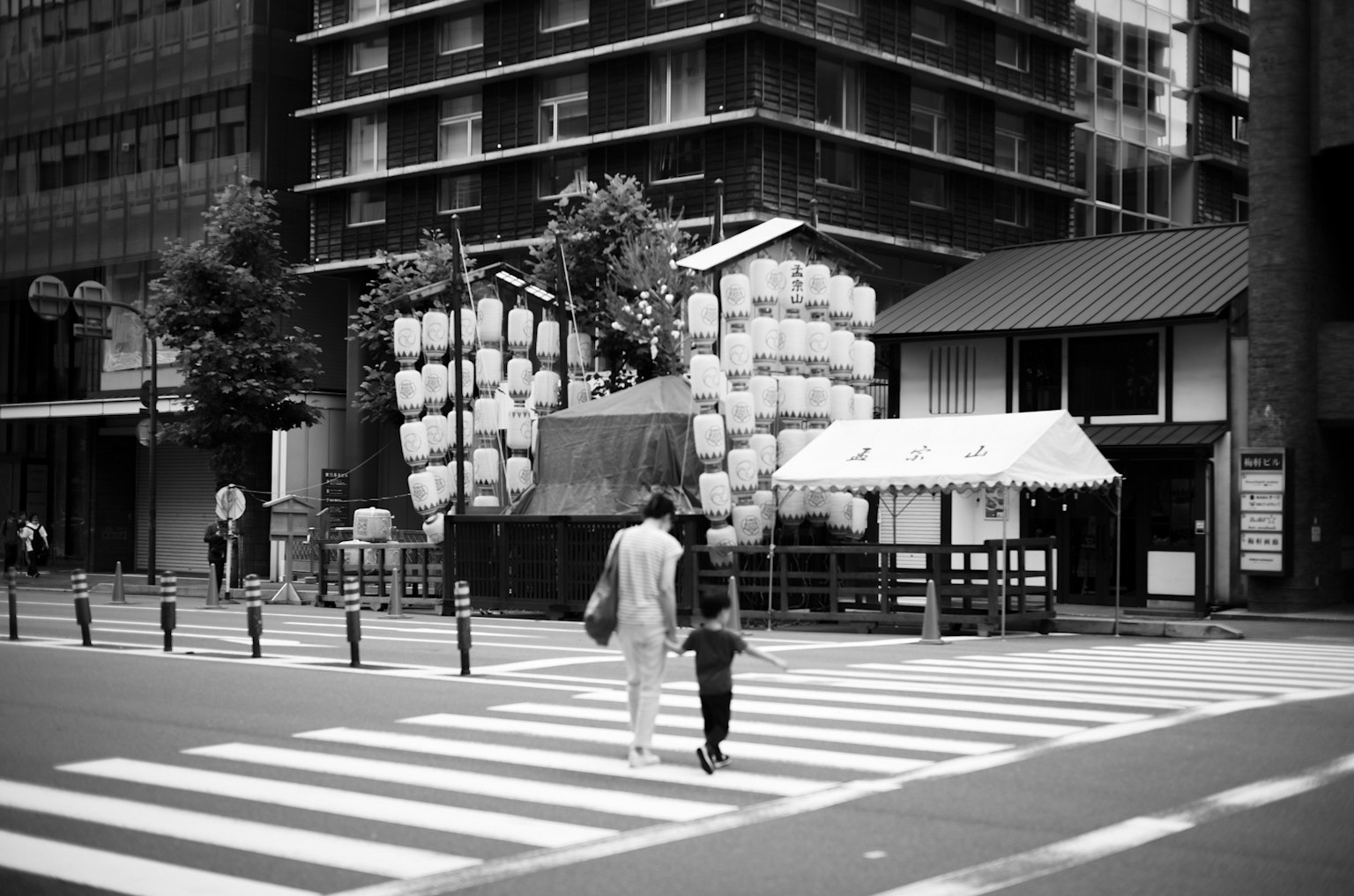 Escena de calle en blanco y negro con un adulto y un niño cruzando un paso peatonal frente a un carro tradicional de festival