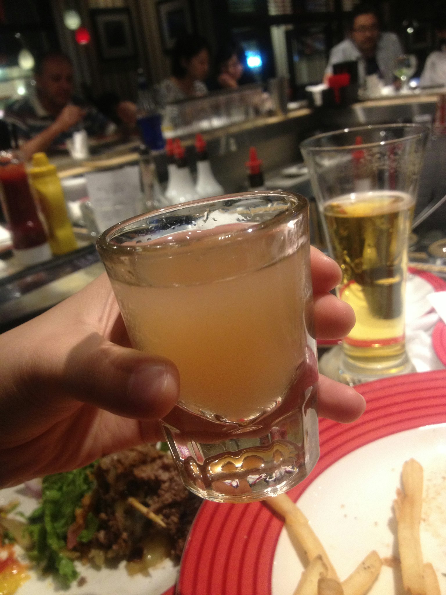 A hand holding a glass of drink with a restaurant background and people
