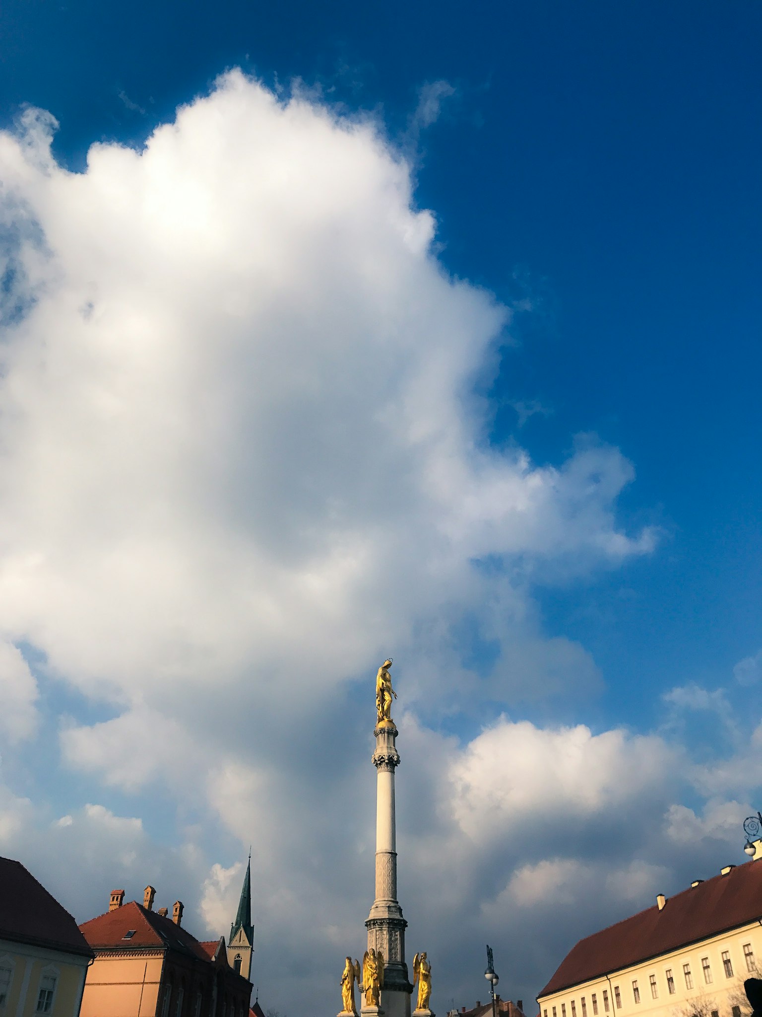 A view of a square with a golden column against a blue sky and white clouds
