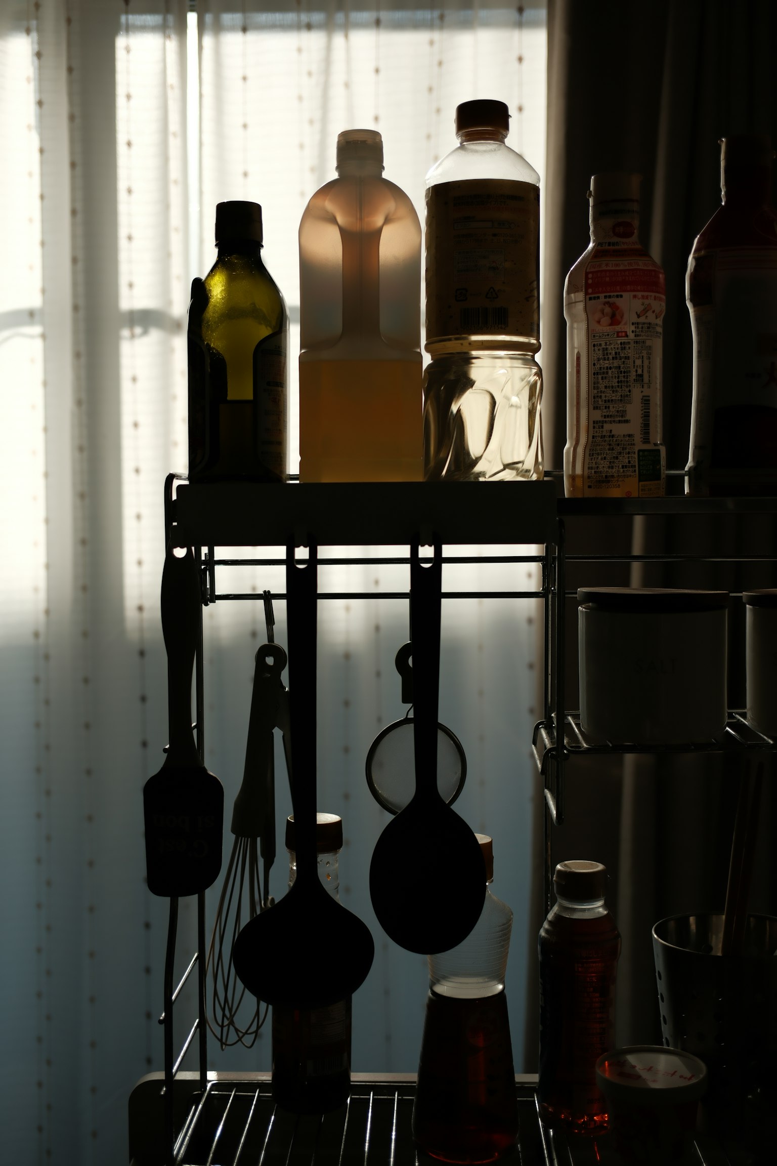 窓の前に置かれた棚に並ぶ調味料の瓶とキッチン器具のシルエット