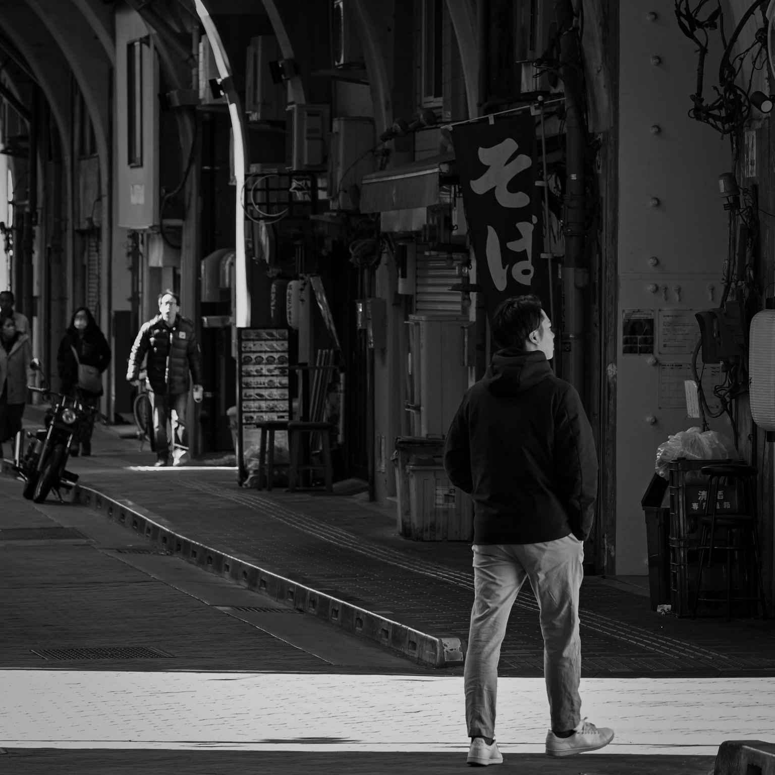 Schwarzweißfoto einer Person, die auf einer Straße mit Geschäften und Schildern im Hintergrund steht