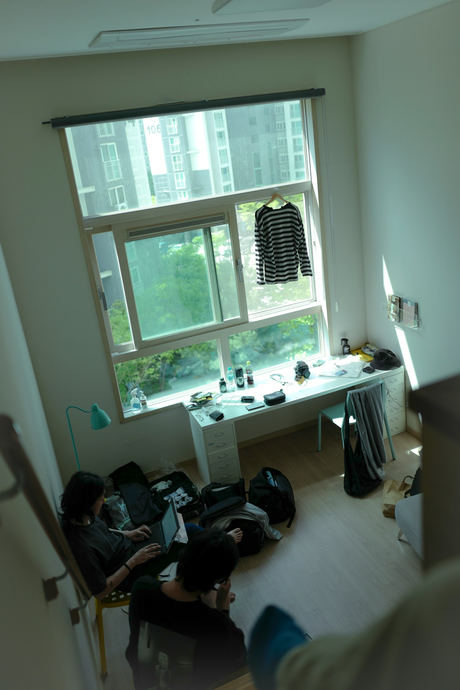 Vista interna di una stanza con una finestra che mostra il verde all'esterno vestiti appesi alla finestra e oggetti sparsi sul tavolo