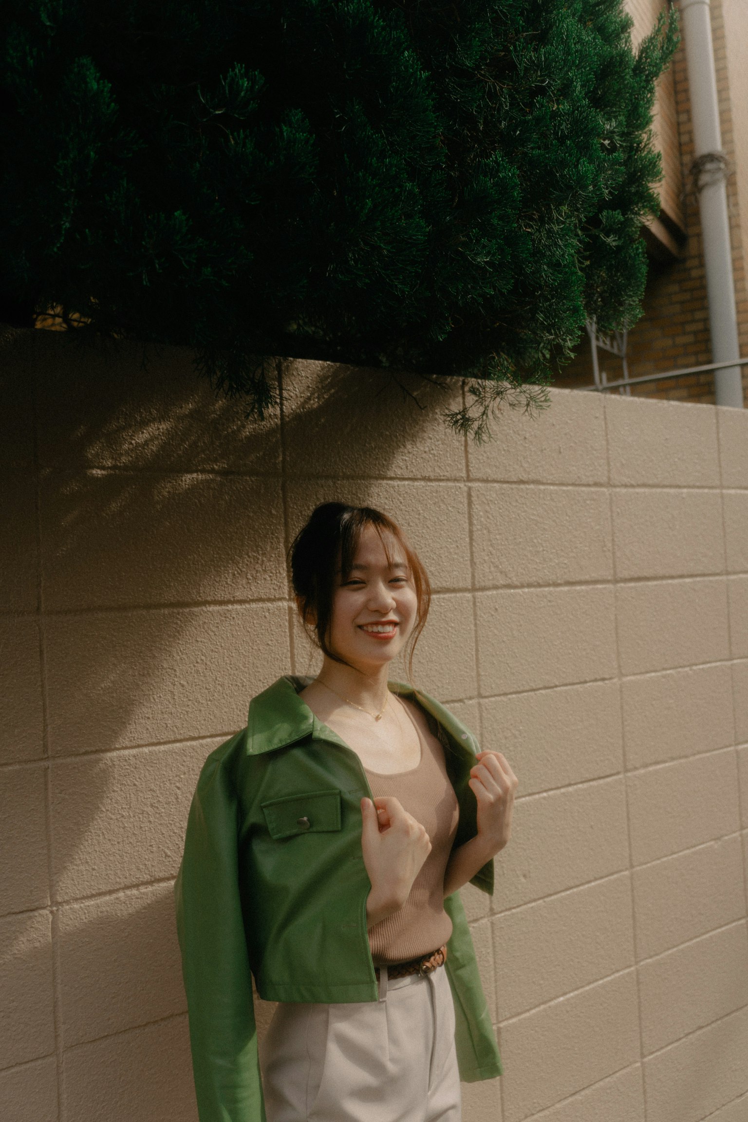 A woman wearing a green jacket smiling against a wall with greenery