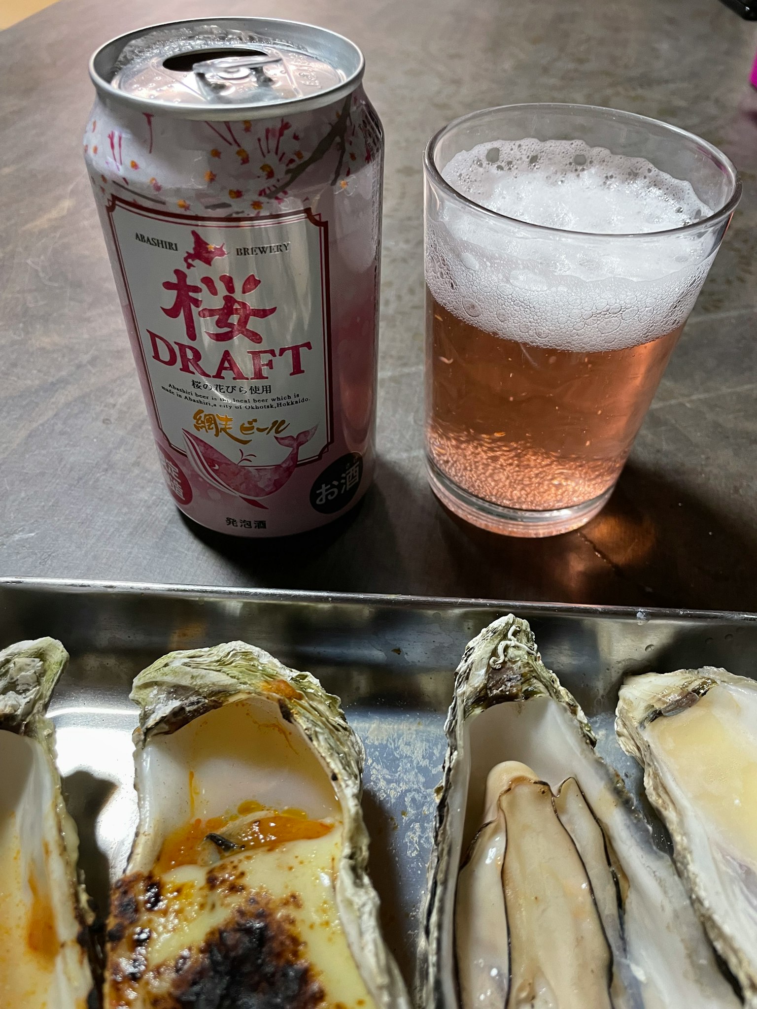 Une canette de bière draft au thème sakura à côté d'un verre de bière mousseuse rose avec des huîtres grillées sur une assiette