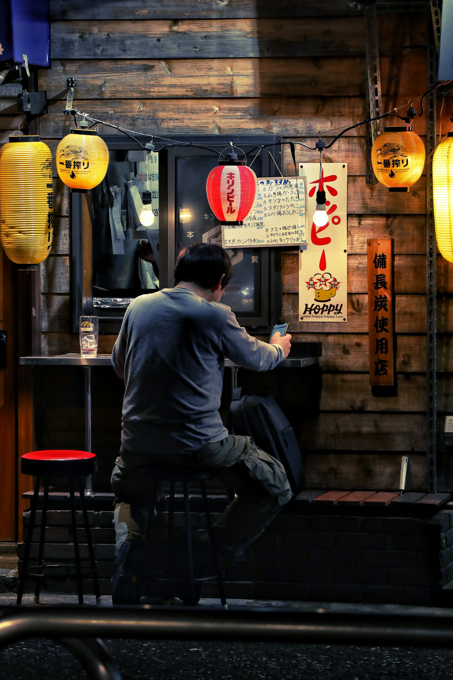 木製の壁の前に座る男性 照明された提灯が周囲を照らす 屋台のメニューが見える