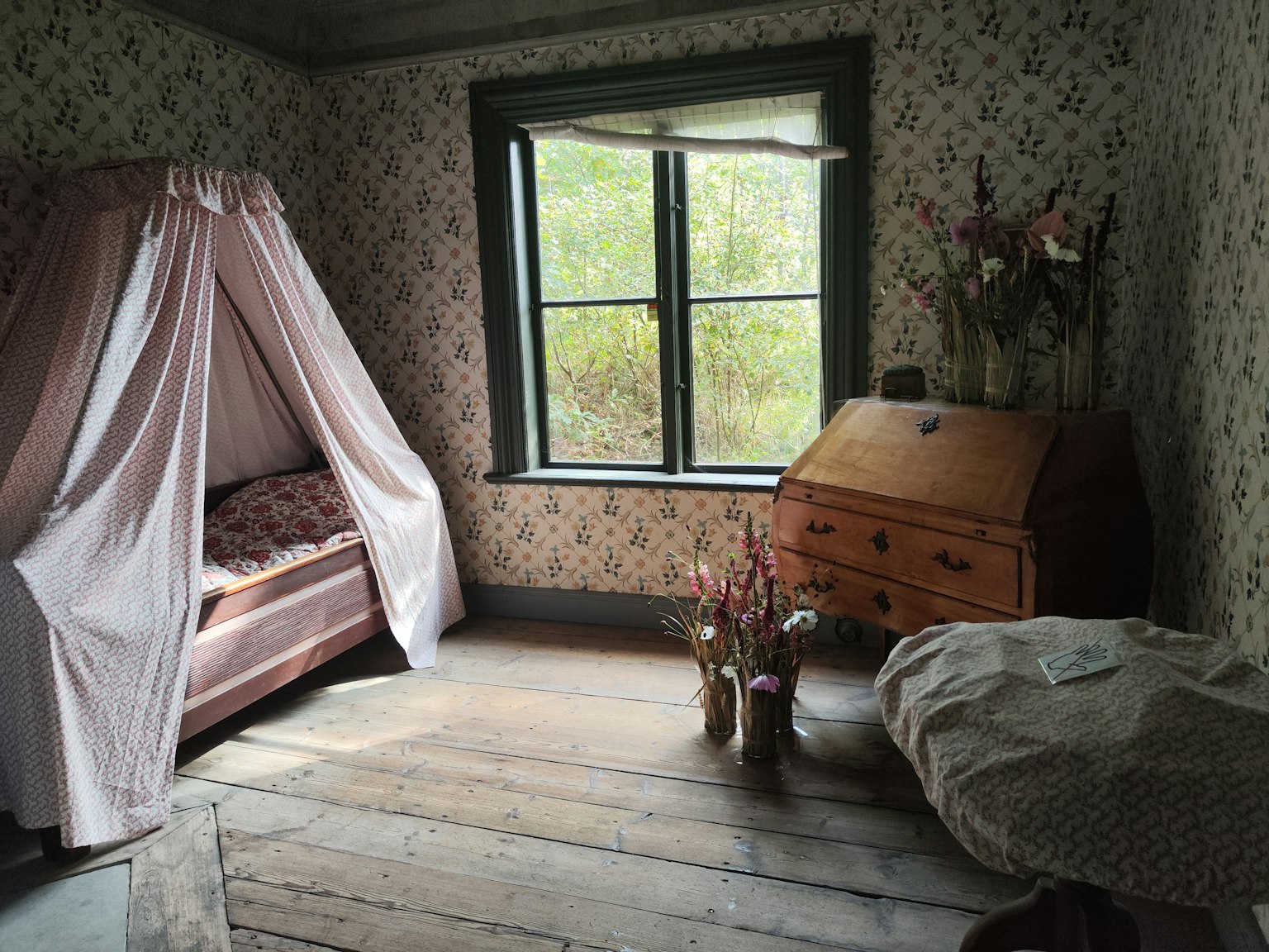 Accogliente camera da letto tradizionale con una finestra luminosa, un letto coperto da tende rosa, mobili in legno e un vaso di fiori