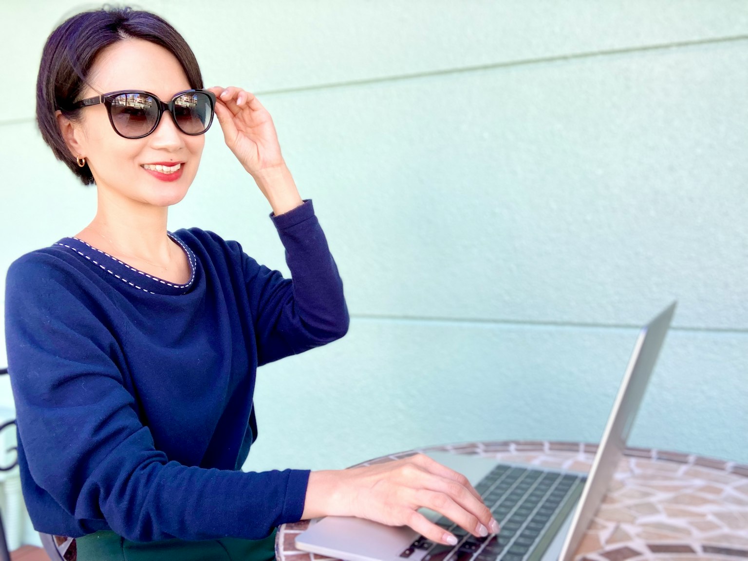 Frau, die draußen an einem Tisch einen Laptop benutzt und lächelt, während sie ihre Sonnenbrille anpasst