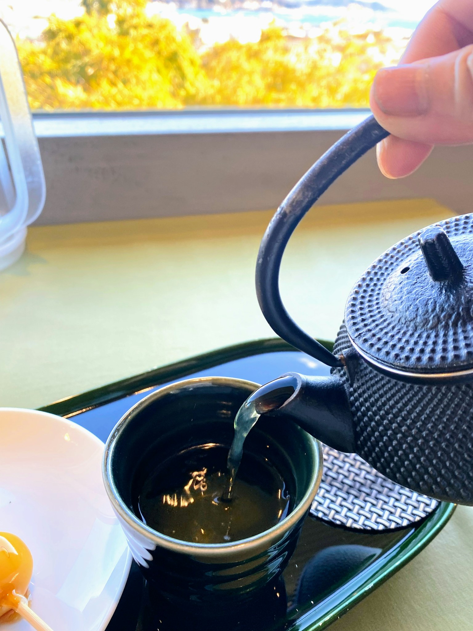 A hand pouring green tea from a cast iron teapot with a natural view outside the window