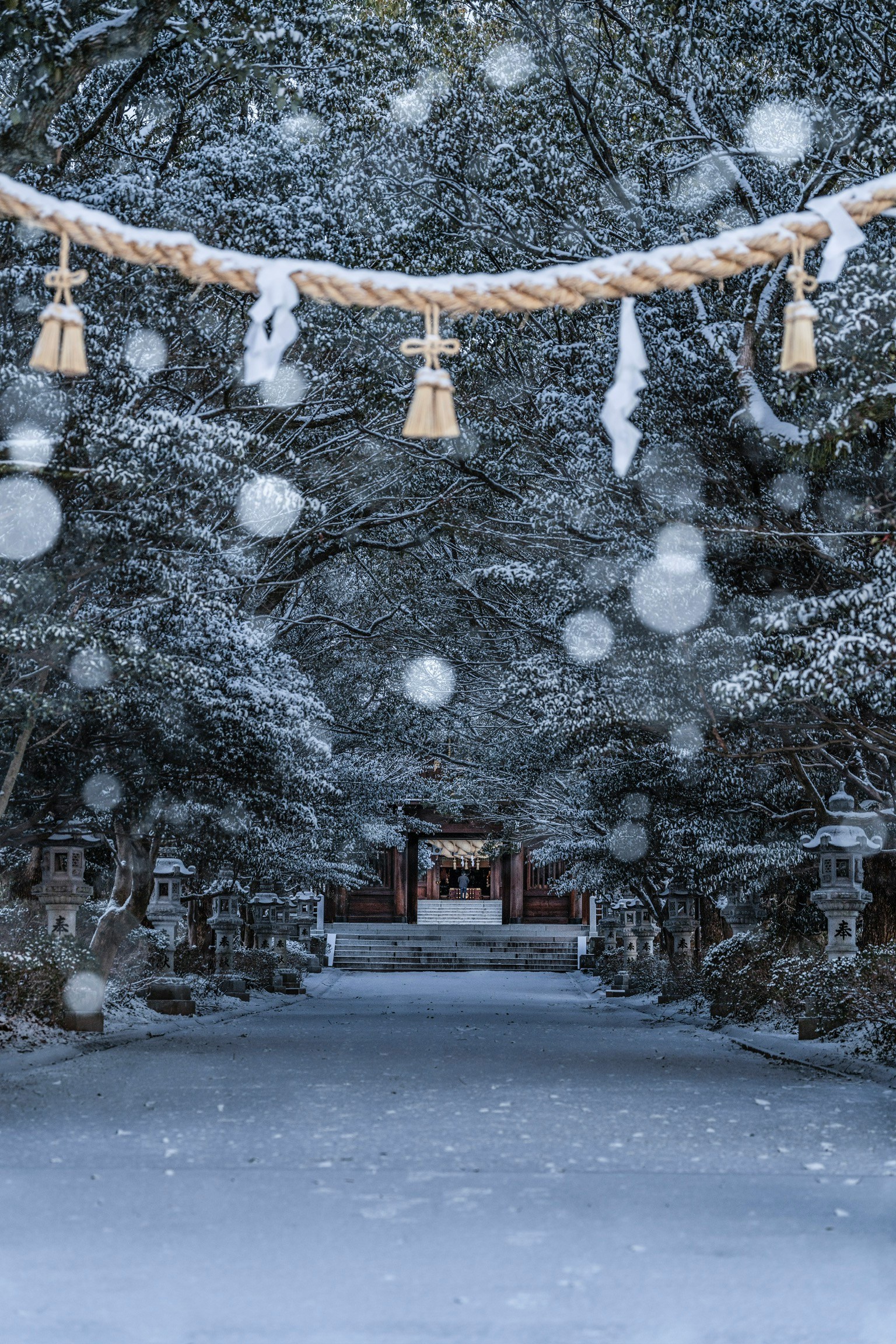雪に覆われた神社の参道に吊るされた注連縄と鈴が見える